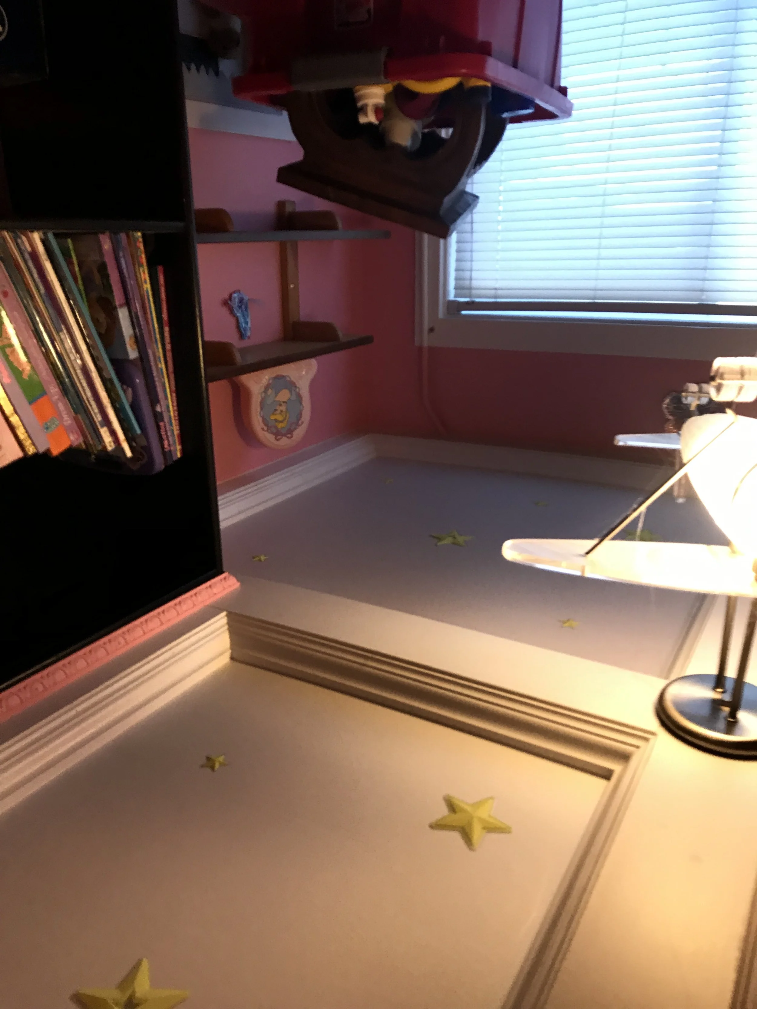 A room with a pink wall, a grey bookshelf with books, a window with closed blinds, yellow star stickers on the carpet, and a wooden desk with a lamp.
