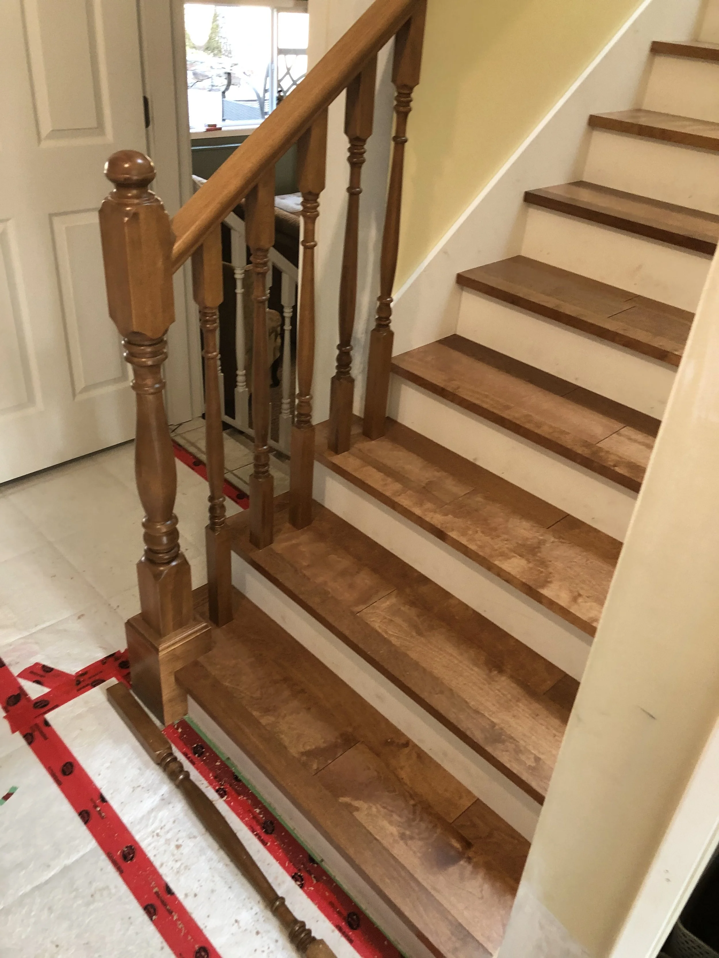 Wooden staircase with a decorative newel post at the bottom, leading upward to an unseen second floor. Light-colored walls and a small section of a window are visible in the background.