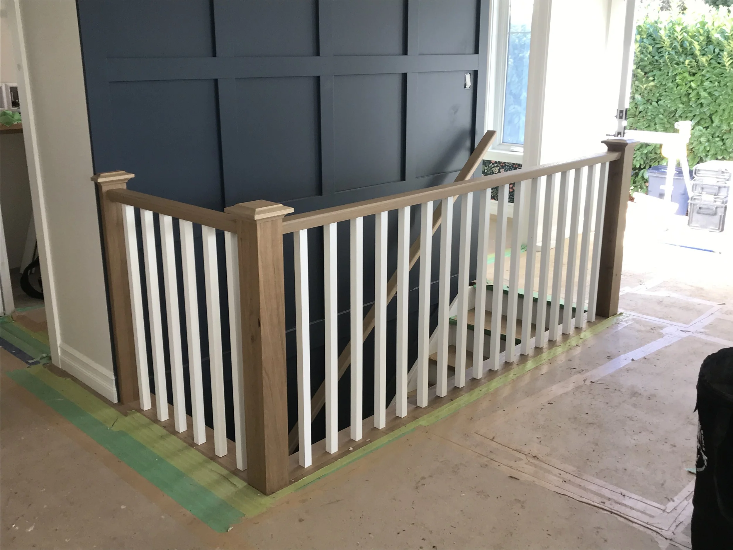 Interior of a house under renovation showing a blue accent wall behind a railing with vertical white and black balusters, with a window to the right allowing natural light, and construction supplies on the floor.