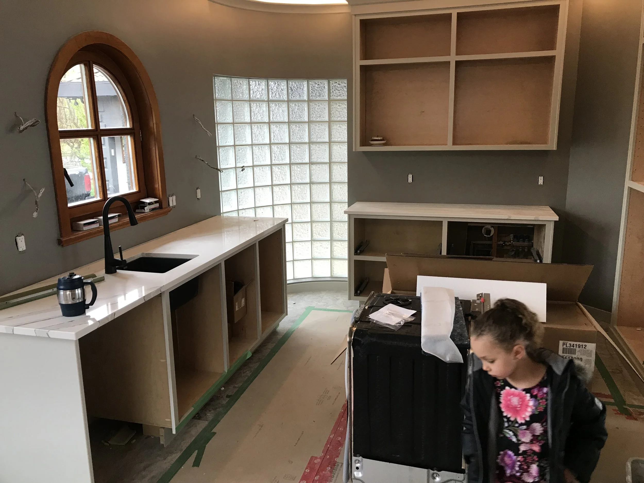 Interior of a house under construction or renovation, with partially installed cabinets, a curved window with wooden trim, glass block window, and a person working on a black appliance or piece of furniture, wearing glasses and a floral dress.