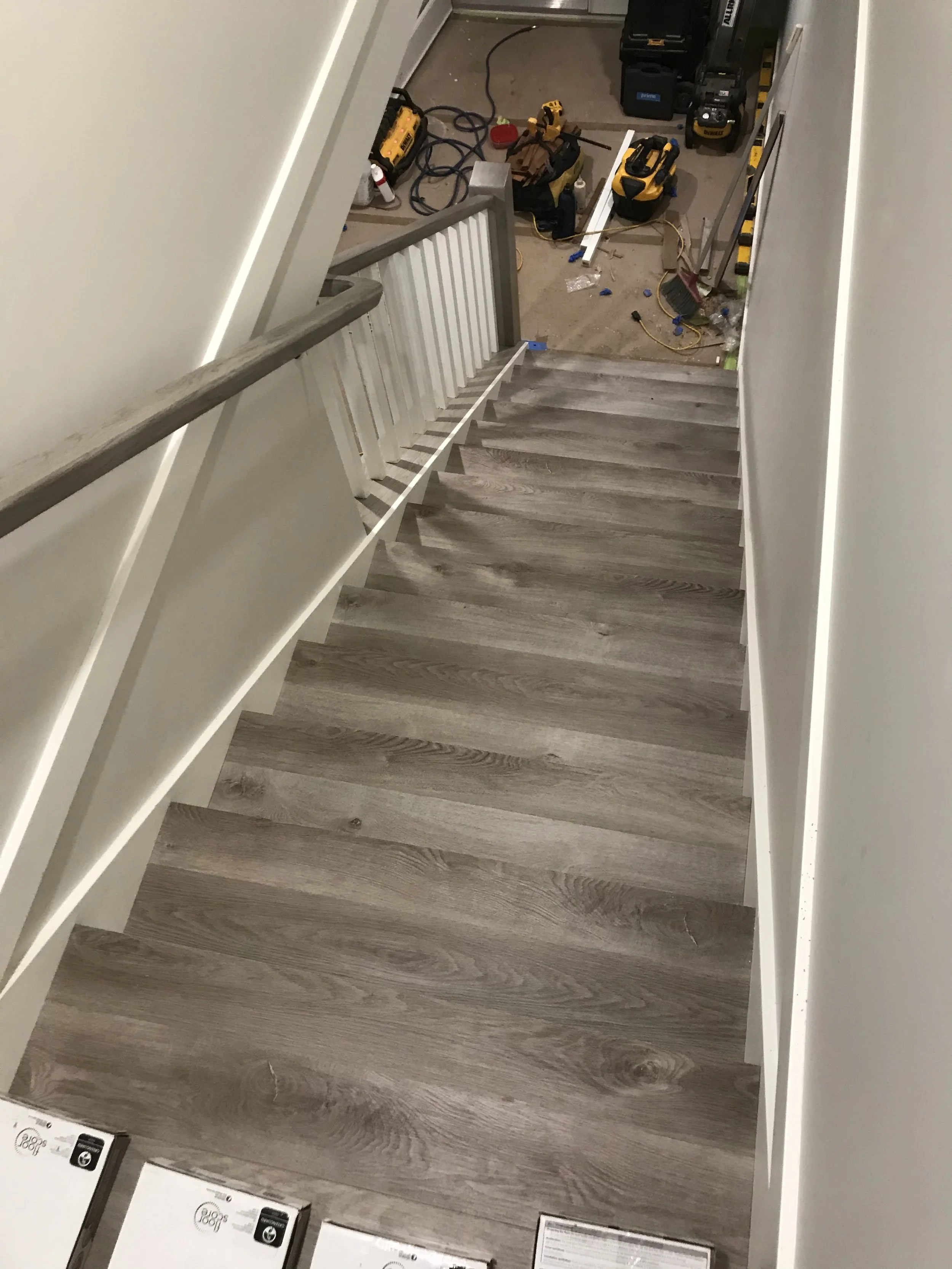View of a staircase with wooden steps, beige walls, and construction tools and materials scattered at the bottom, indicating ongoing renovation.
