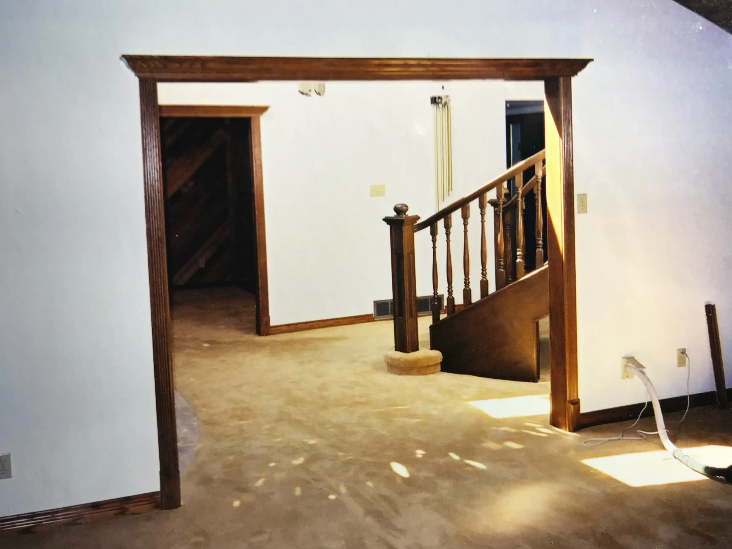 Living room with wooden staircase, doorway, and white walls, illuminated by warm lighting, with a white fan plugged into an outlet on the right.