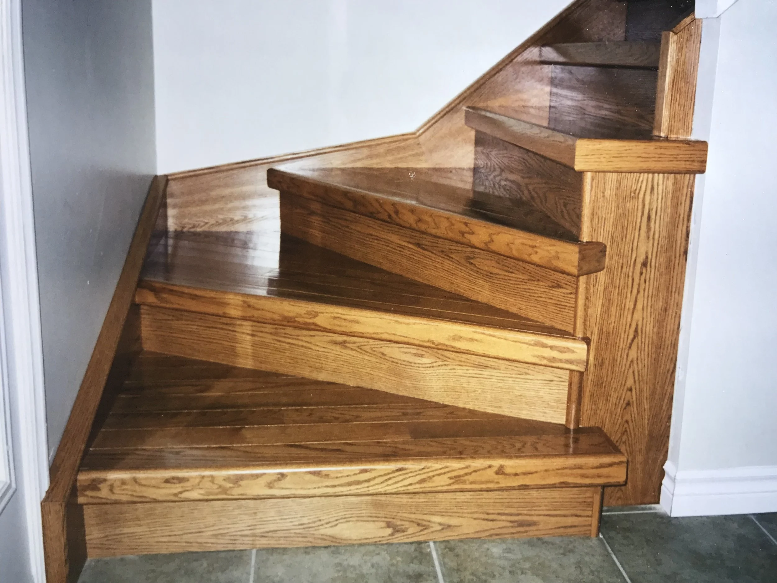 Wooden staircase with three steps and a handrail on the right side, next to a gray wall and tiled floor.