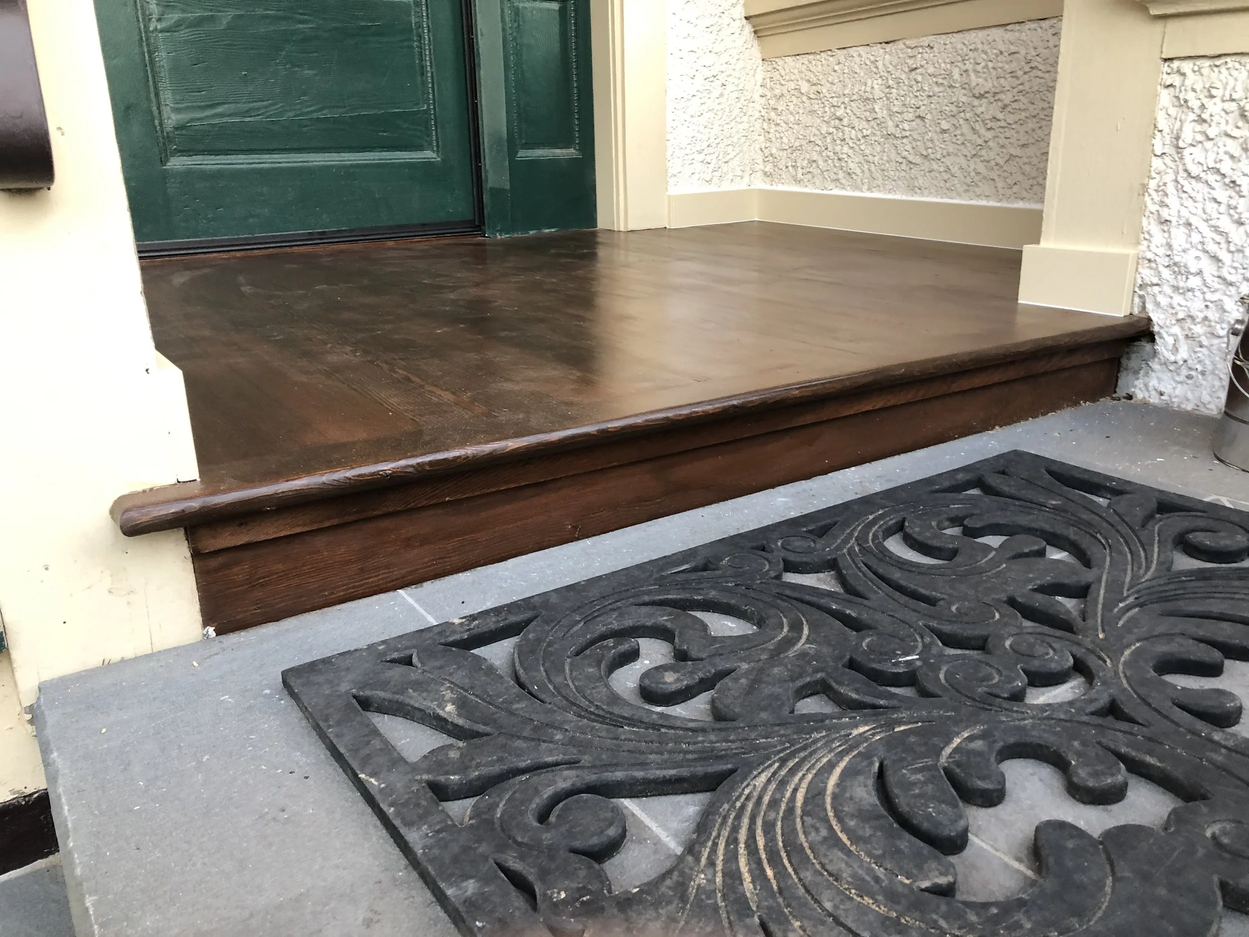 The image shows a front porch area with a wooden step and two black decorative mats. The porch has a textured wall and a green door.