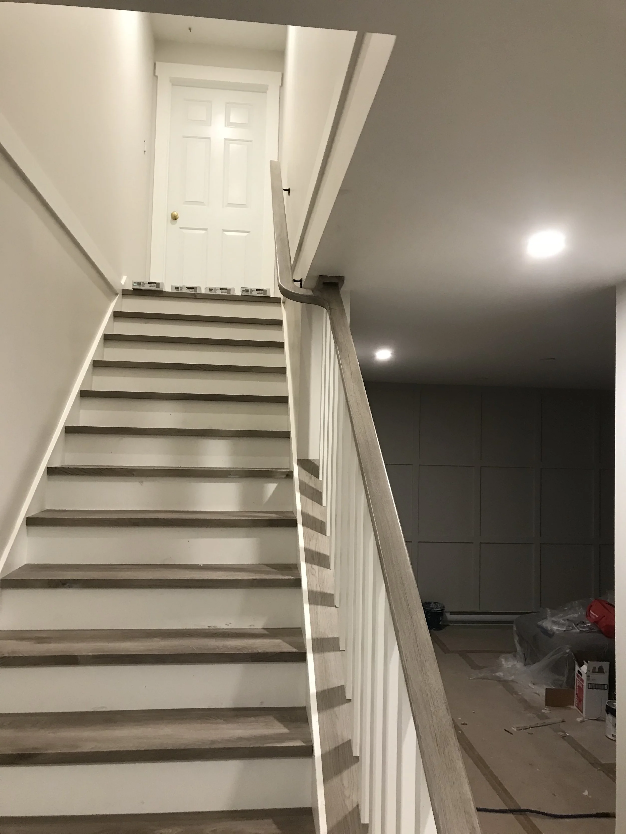 Photo of a staircase with a white door at the top, male handrail, and beige walls, with construction supplies and debris at the bottom.