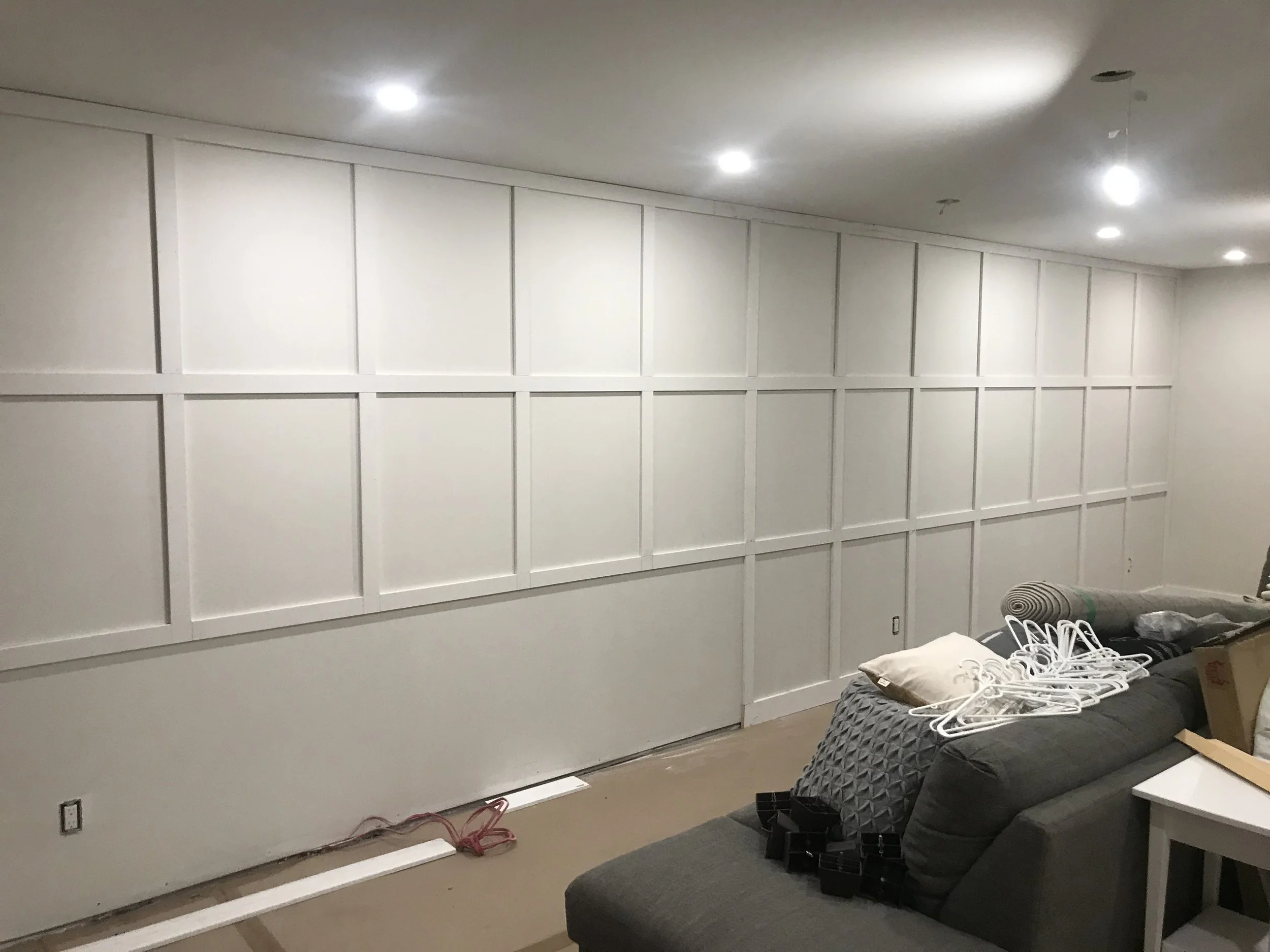 A partially constructed room with white paneled walls and a white ceiling, with several recessed lights. There is a black couch with items, including a white pillow, some hangers, and a cardboard box, and a small white table.