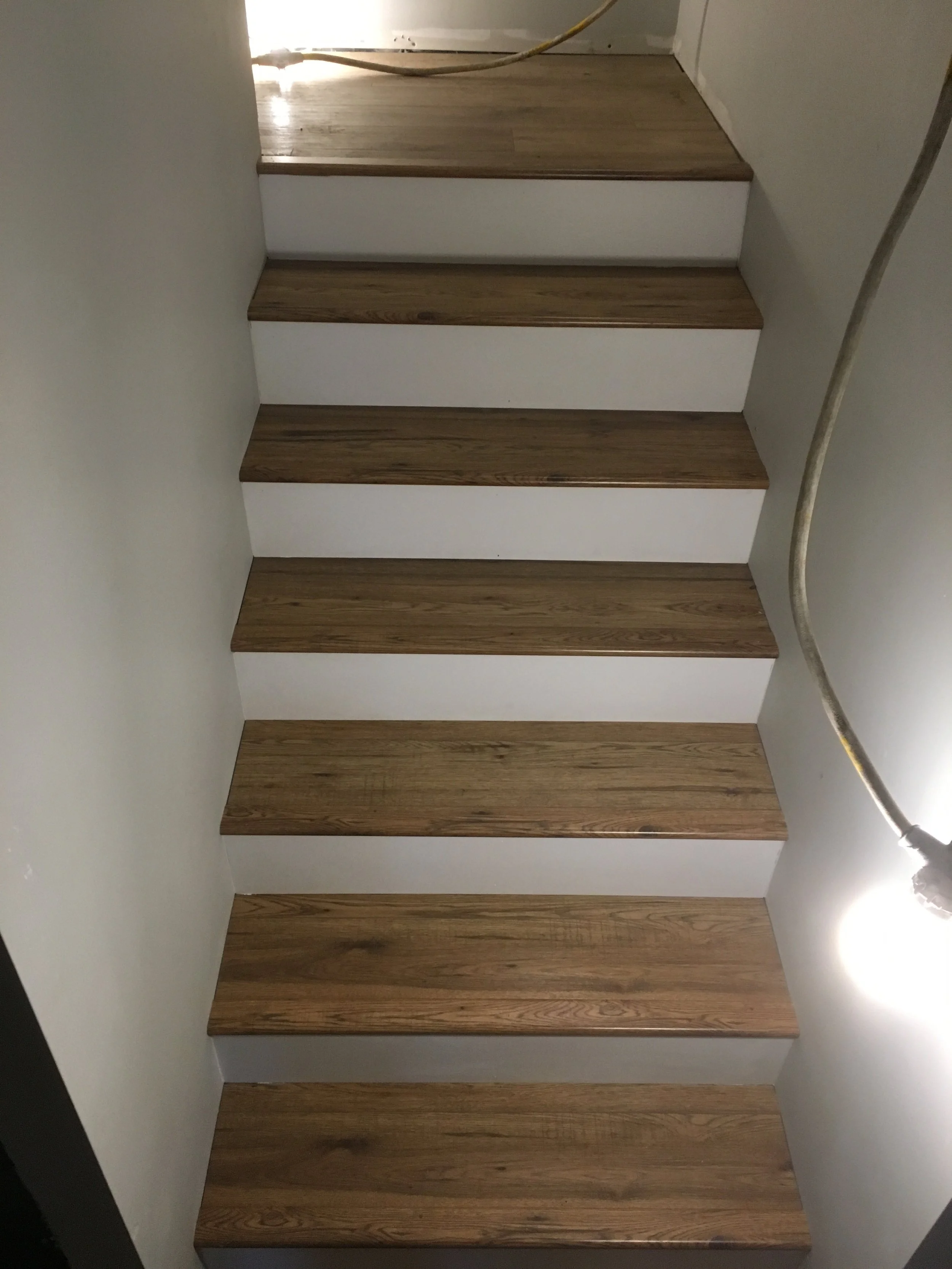 A staircase with five wooden steps and white risers, with a white wall on the left and a metal handrail on the right side.