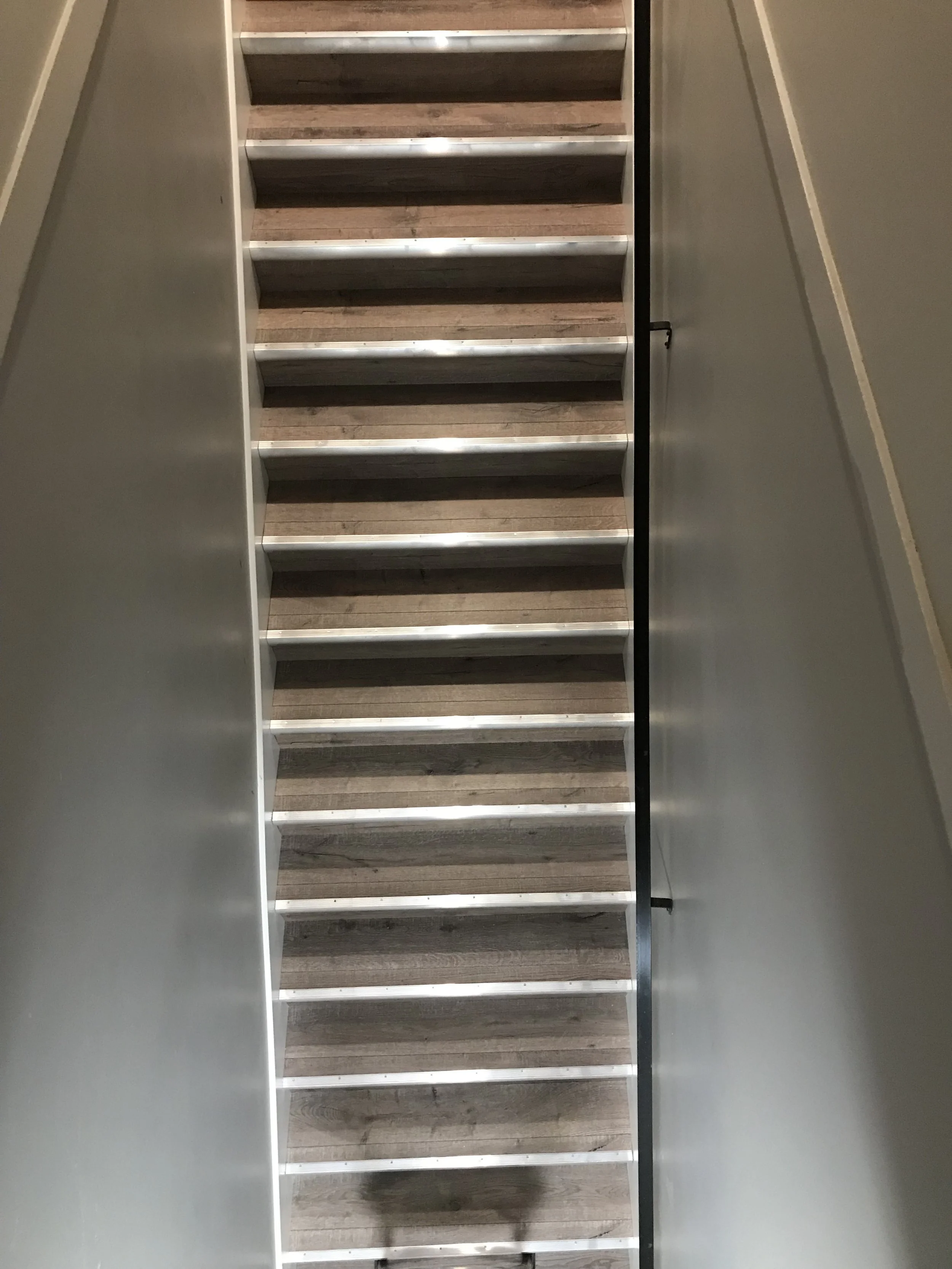 A photo of a staircase with wooden steps and metal edges, viewed from the bottom looking up, with beige walls on either side.