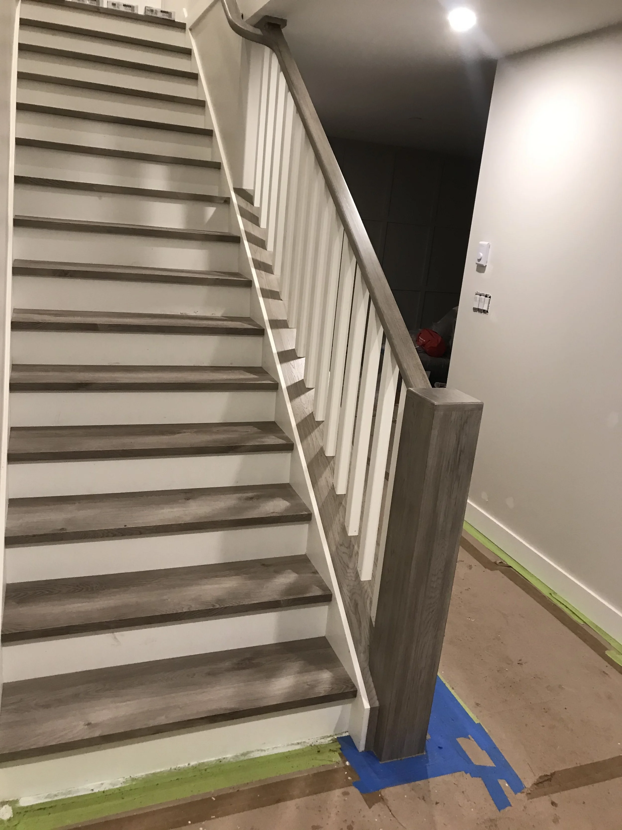 Inside view of a staircase with wooden steps, white risers, a wooden handrail, and white balusters, with a wall in the background and painter's tape on the floor.