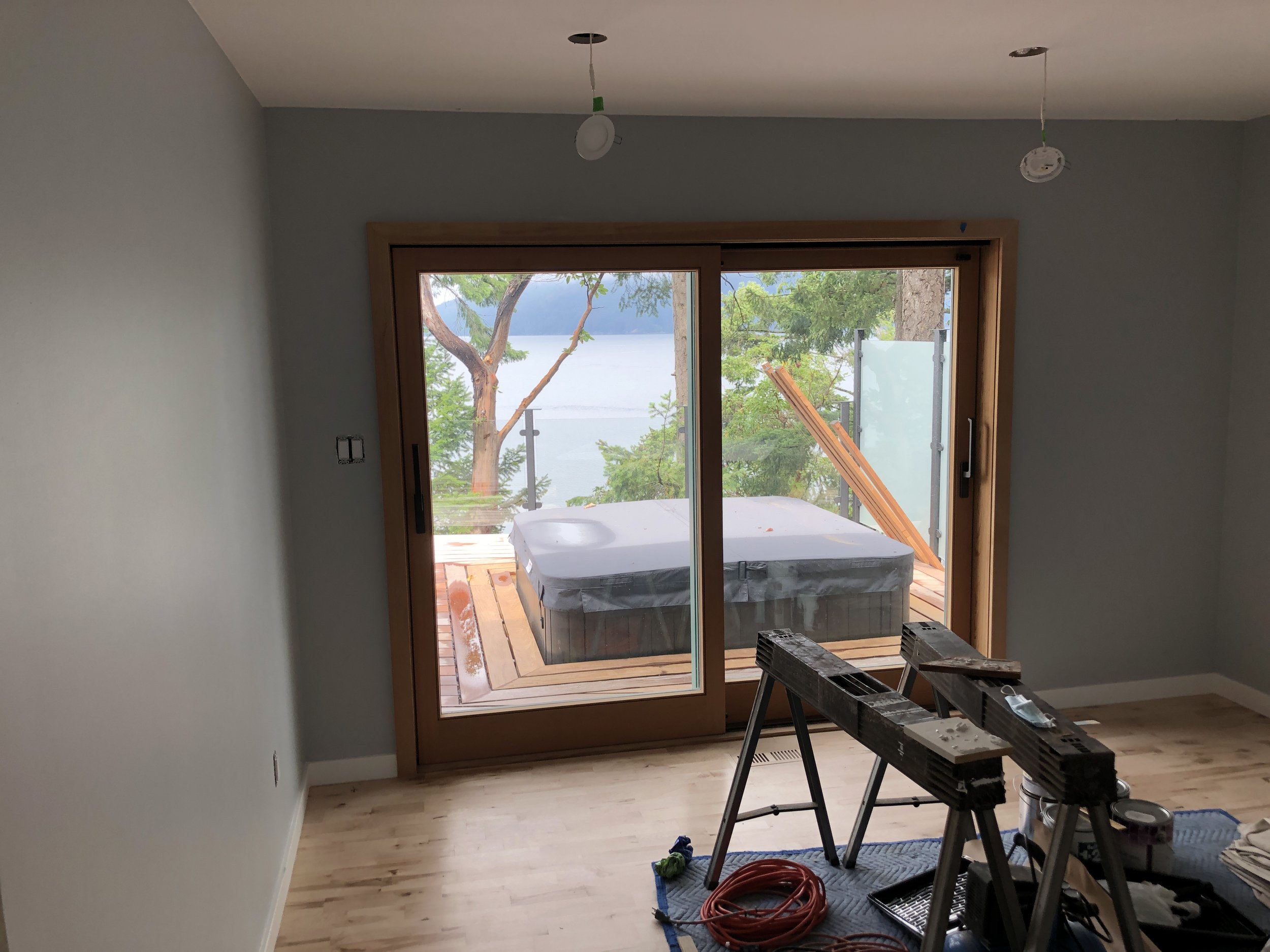 Interior of a room under construction with sliding glass doors leading to an outdoor deck overlooking a lake and trees. Construction tools are on the floor and a ladder is inside the room.