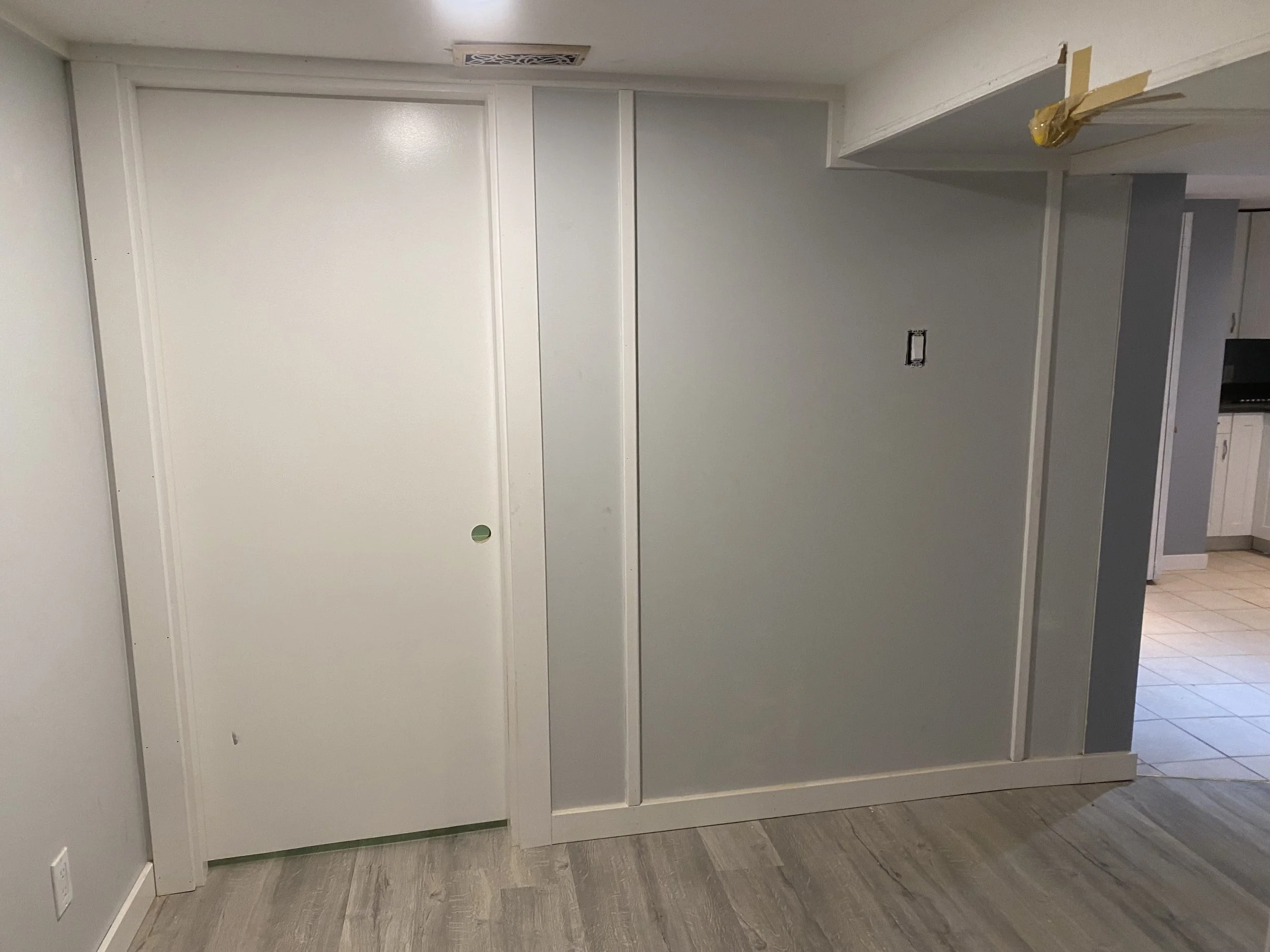 Interior of a room with white walls, a white door, and a floor with wood-like tiles. There is a section of the ceiling with a vent and a corner with taped-seam. The room appears to be under construction or renovation.