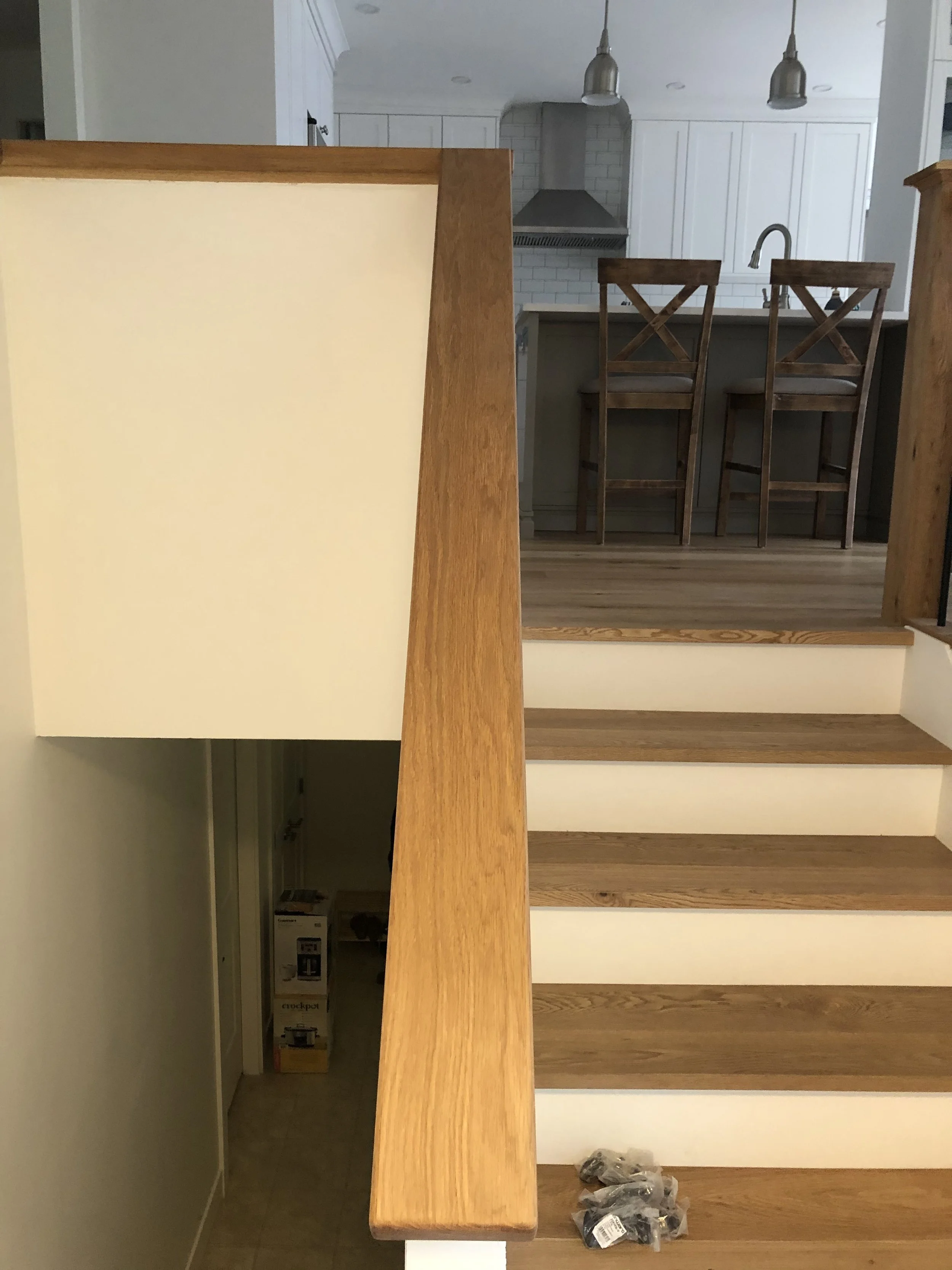 Kitchen with a wooden counter, white cabinets, and a white tiled wall. Two wooden chairs are in front of the counter, and there are pendant lights hanging from the ceiling. Items are on the floor near the wall, including plastic bags with tools or su