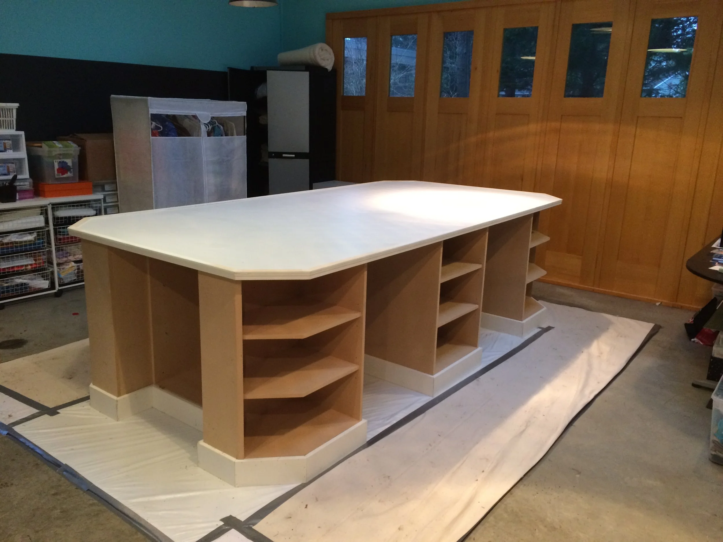 Interior of a room with a wooden wall and large windows, featuring an unfinished kitchen island or counter with open shelves, and storage containers and shelves on the side.
