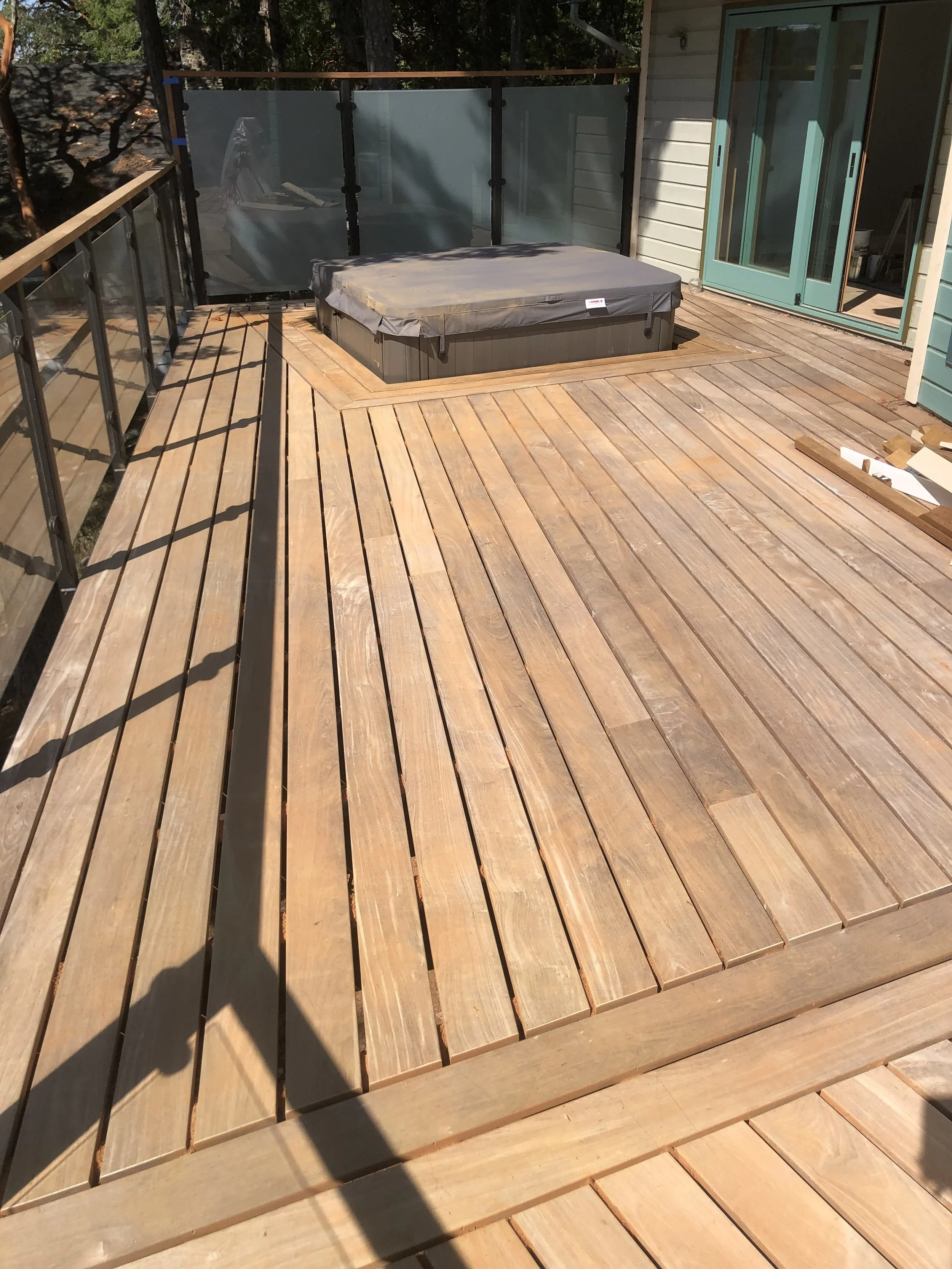 A wooden deck with a hot tub covered by a grey cover, surrounded by a black railing with glass panels. There is a house with blue sliding doors and a white siding wall. Shadows of trees and a person are visible on the deck.
