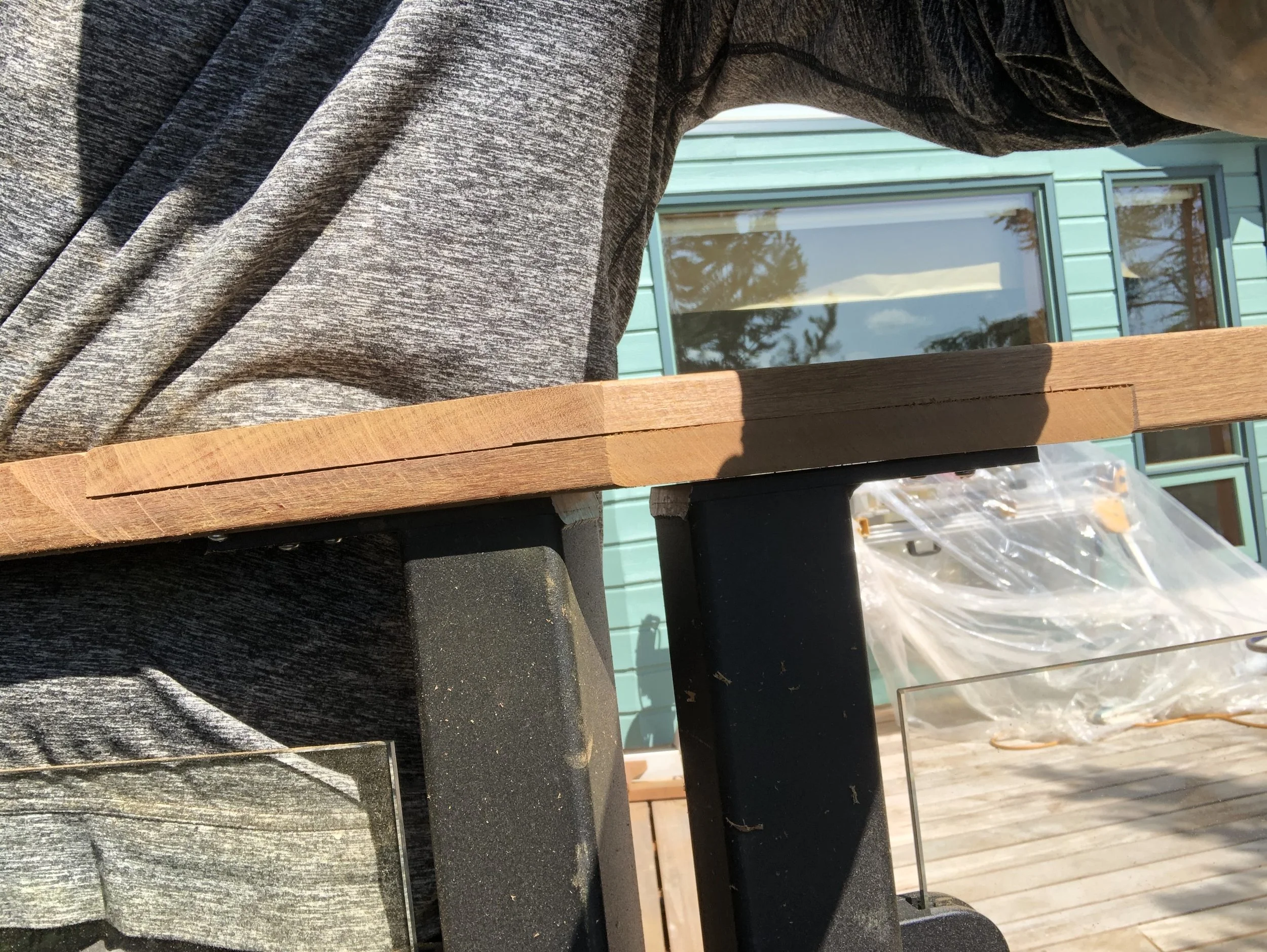 Close-up of a wooden table leg and part of a person wearing gray, textured sweatpants, with a residential house with green siding and windows reflected in the background.