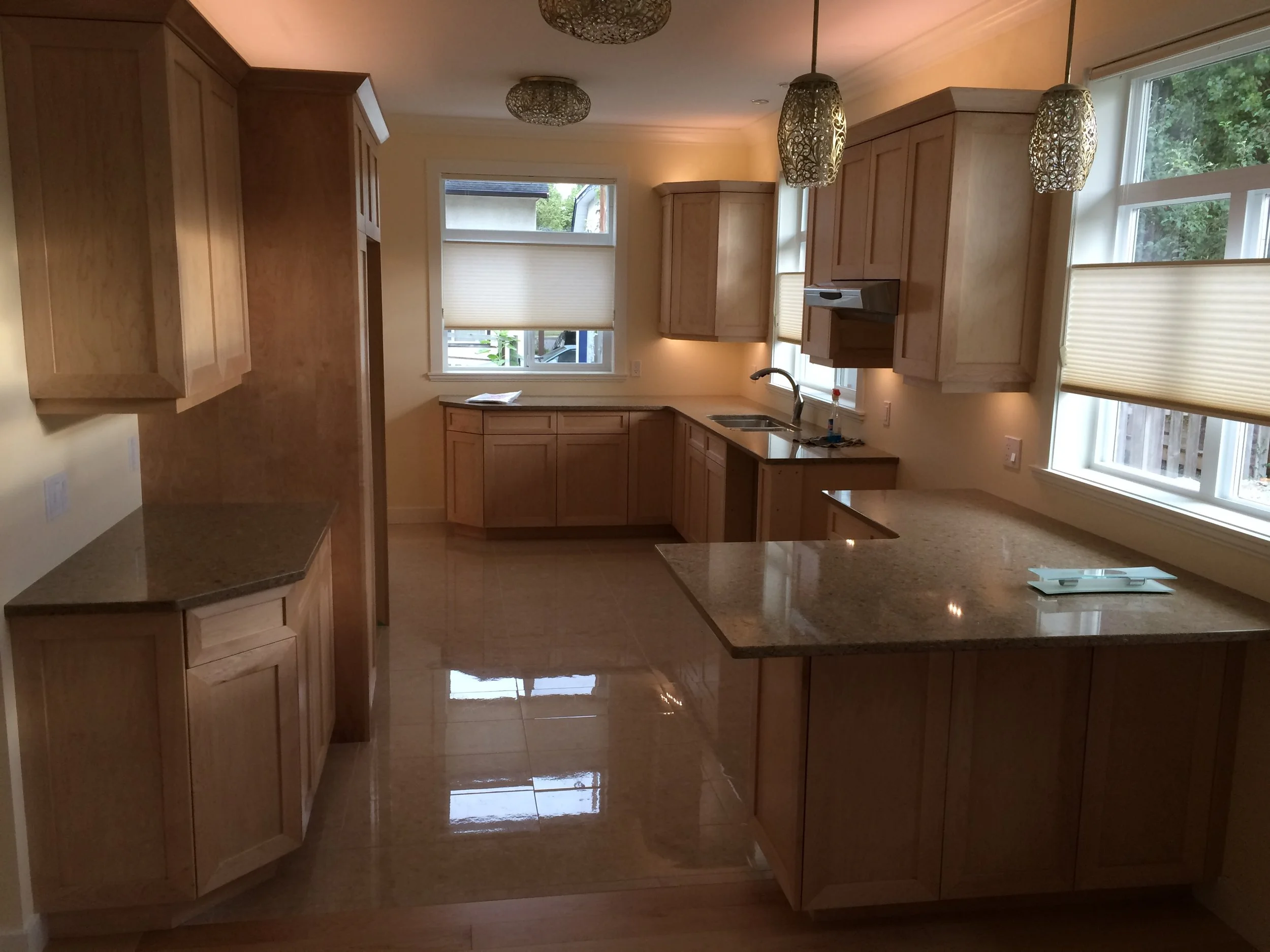 A kitchen with wooden cabinets, granite countertops, a double sink, windows with blinds, and pendant lights hanging from the ceiling.