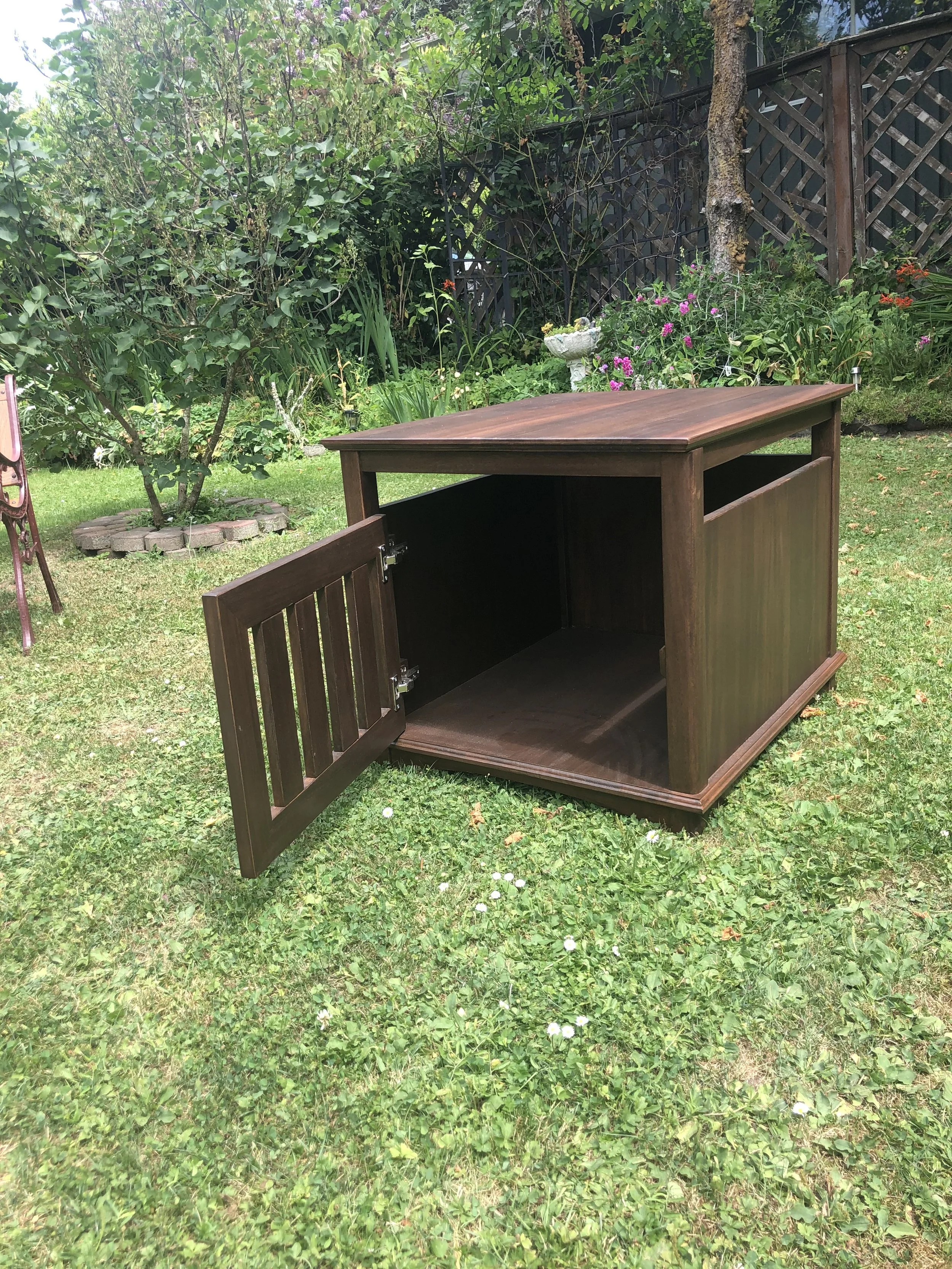 A garden with lush green grass, various flowering plants, and trees. There is a small statue or birdbath in the background. In the foreground, an open wooden cabinet with a slat design on the side.