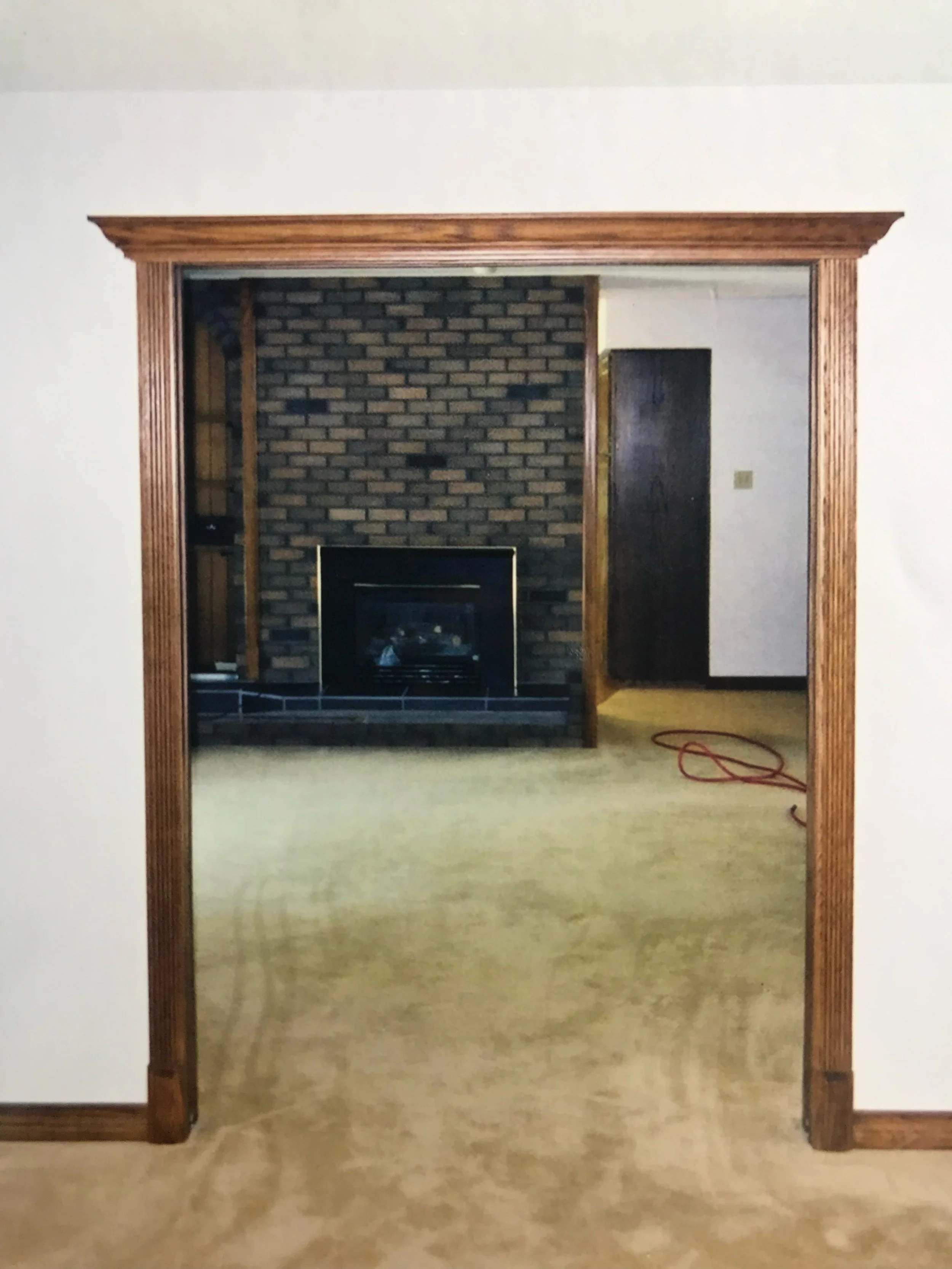 Photo of a doorway with a wooden frame leading into a room with a brick fireplace and a brass trim. The room has light-colored walls and a beige carpet.