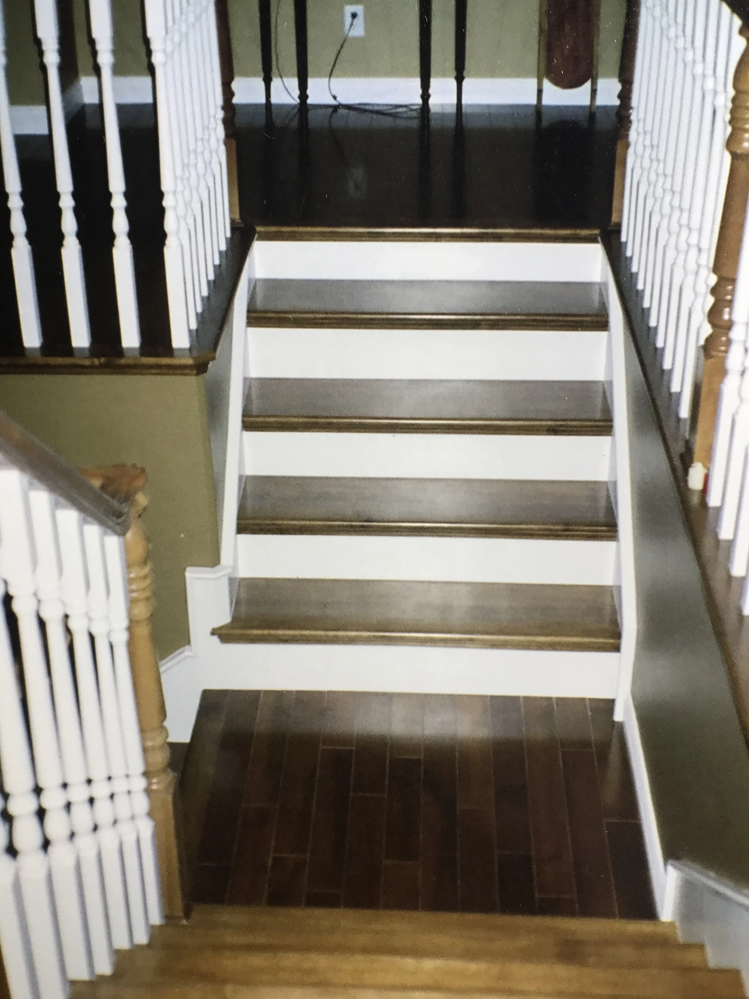 Image of a staircase with wooden steps and dark-colored treads, flanked by white and wooden spindled railings on both sides. A portion of hardwood flooring is visible at the bottom of the stairs, and an electrical outlet with a plugged-in cord is vis