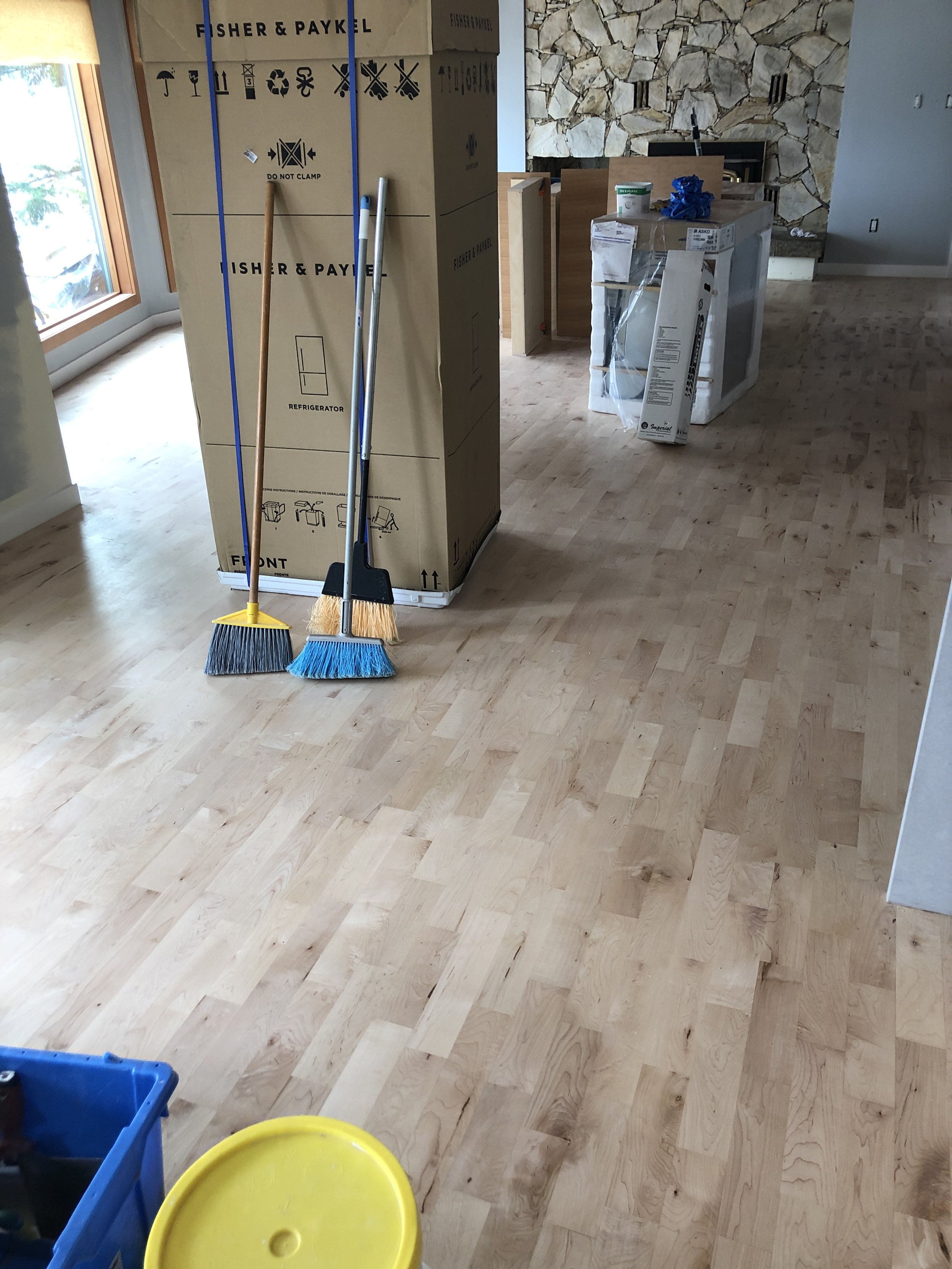 Interior of a house under renovation with an empty wooden floor, a large cardboard box labeled 'Fisher & Paykel,' cleaning tools such as a broom and dustpan, and various construction materials and boxes.