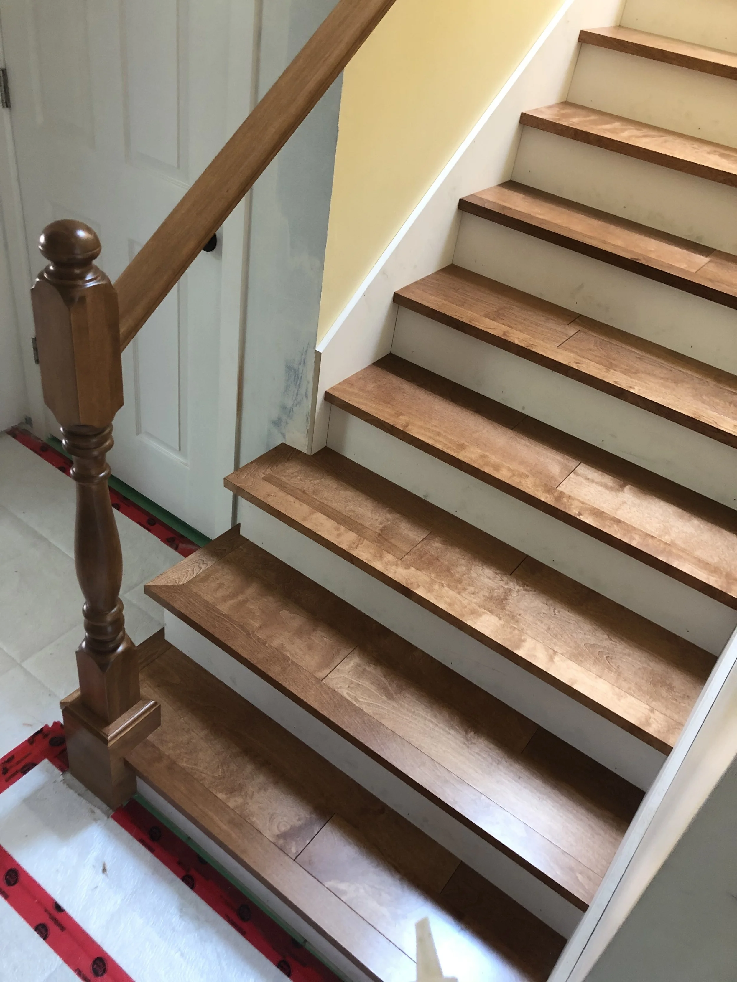 Photo of wooden stairs with a brown handrail, showing the edge of the stairs and part of a yellow wall next to them.