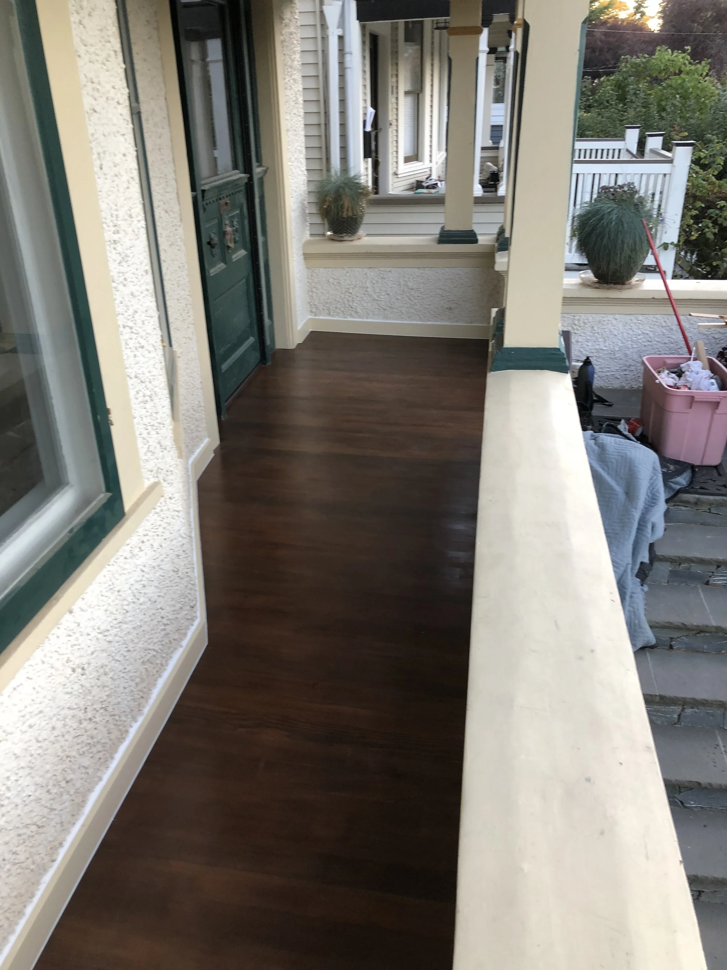 A porch area with brown wooden flooring, a green door with mail slots, multiple windows, a small potted plant, a larger potted plant, and a pink box with clutter. The porch is partially covered with white railings and decorative columns. There are tr