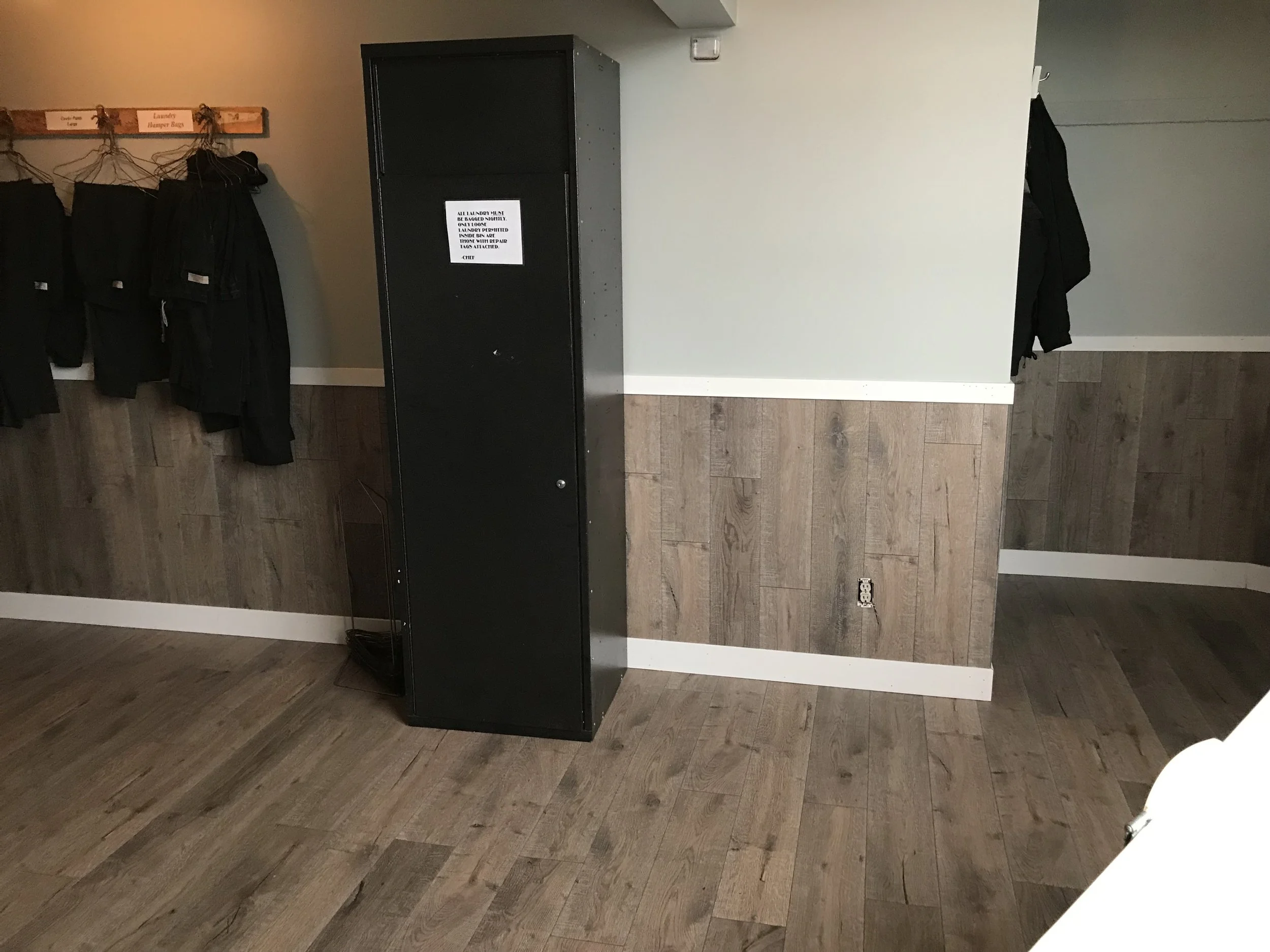 A black luggage locker with a note attached to it, a small area has black hangers with black coats hanging, and wooden flooring with a wall in the background.