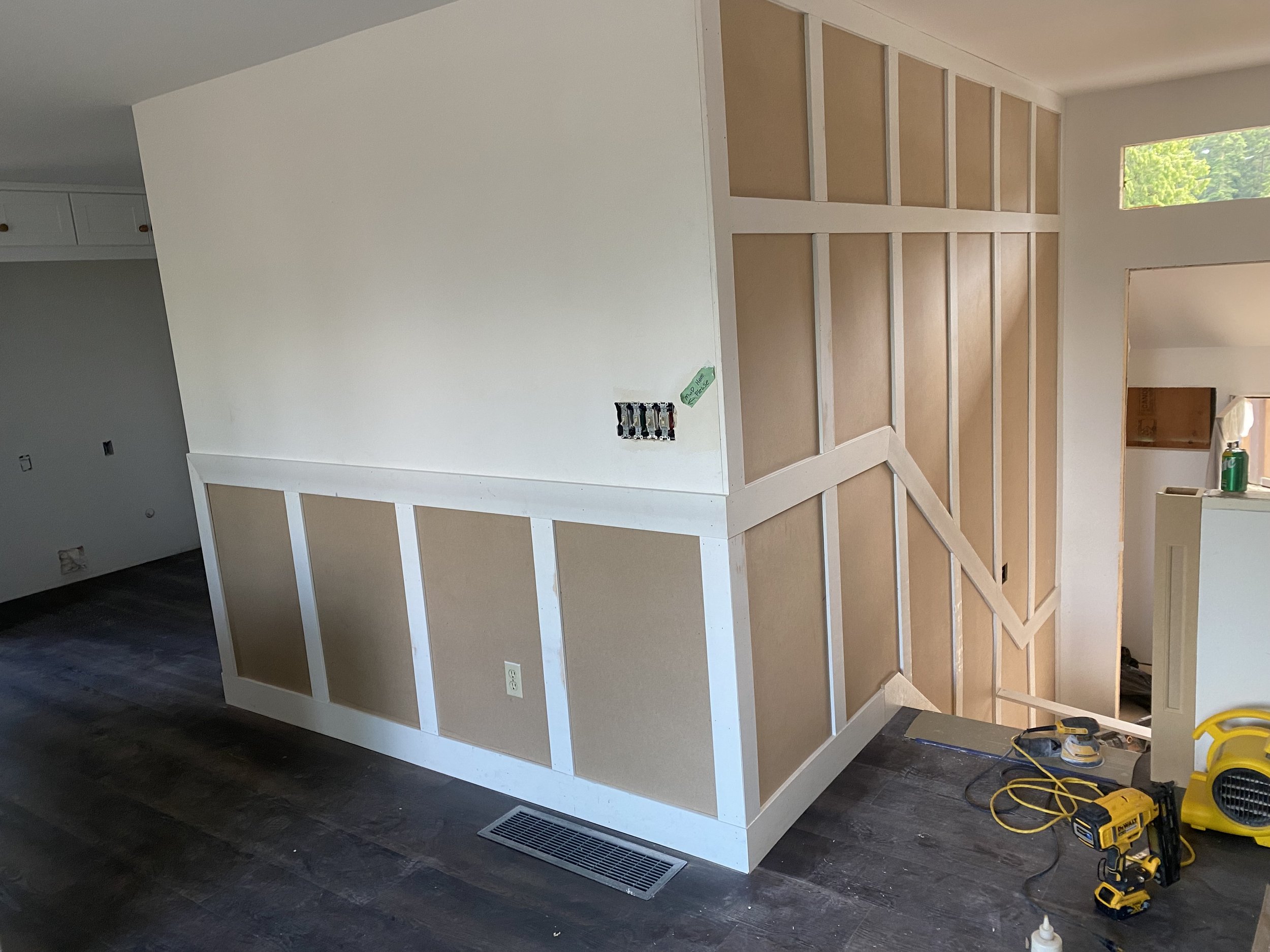 Interior of a house under construction with a partially built wall and framing on top, tools and electrical wires on the floor, and a window showing a view of green trees outside.
