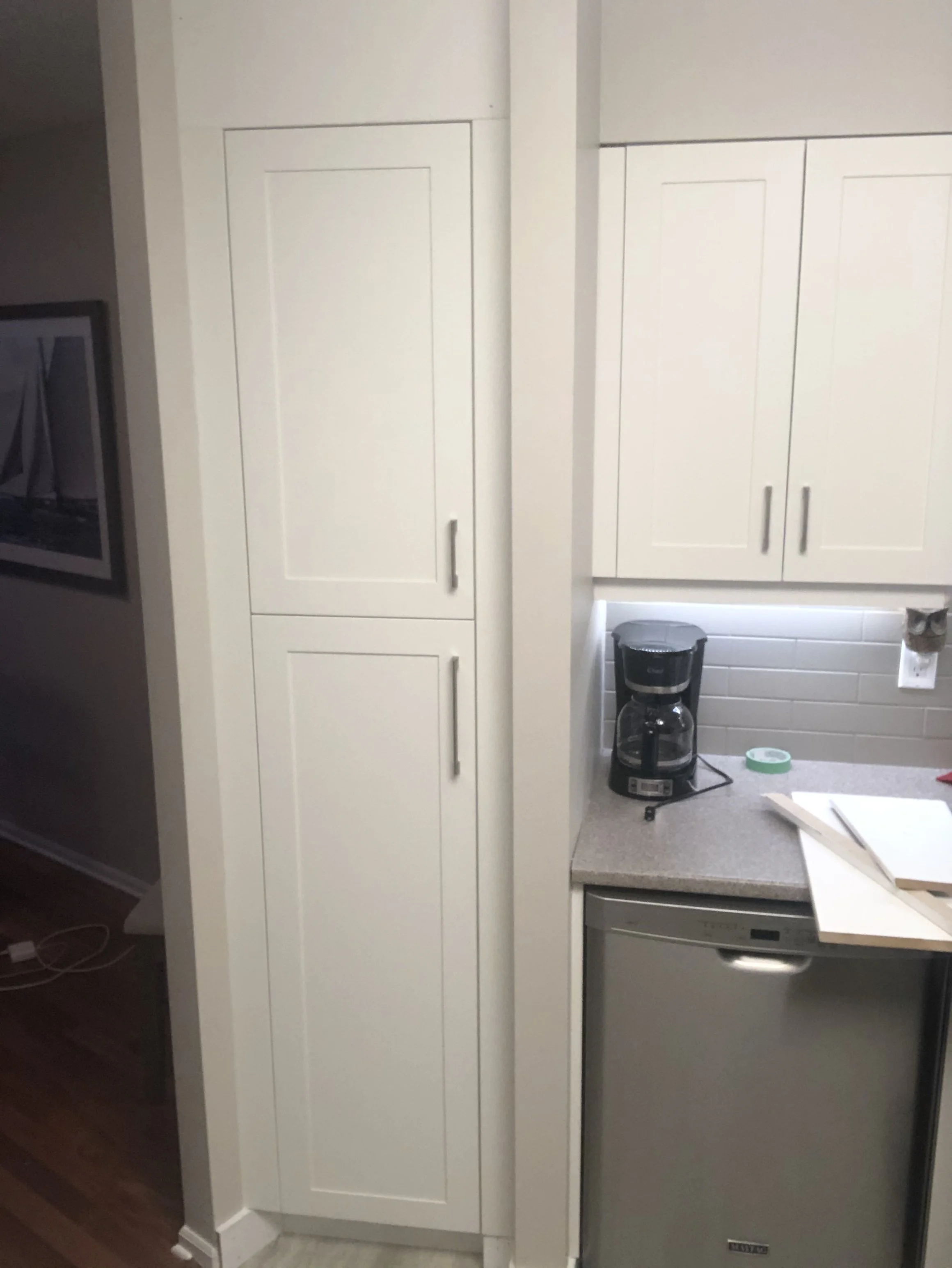 A kitchen corner with white cabinets, a coffee maker on the countertop, a roll of tape, and a piece of paper or wood on the counter. A dishwasher is visible below the counter.