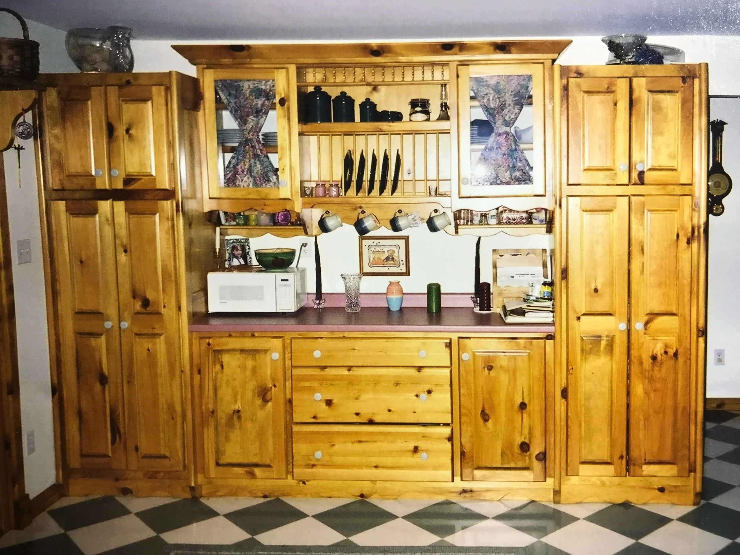 A kitchen with wooden cabinets and a pink countertop. The cabinets have white knobs, and there are decorative curtains on the windows behind the cabinets. There are various kitchen items such as cups, jars, and a framed photo on the wall and countert