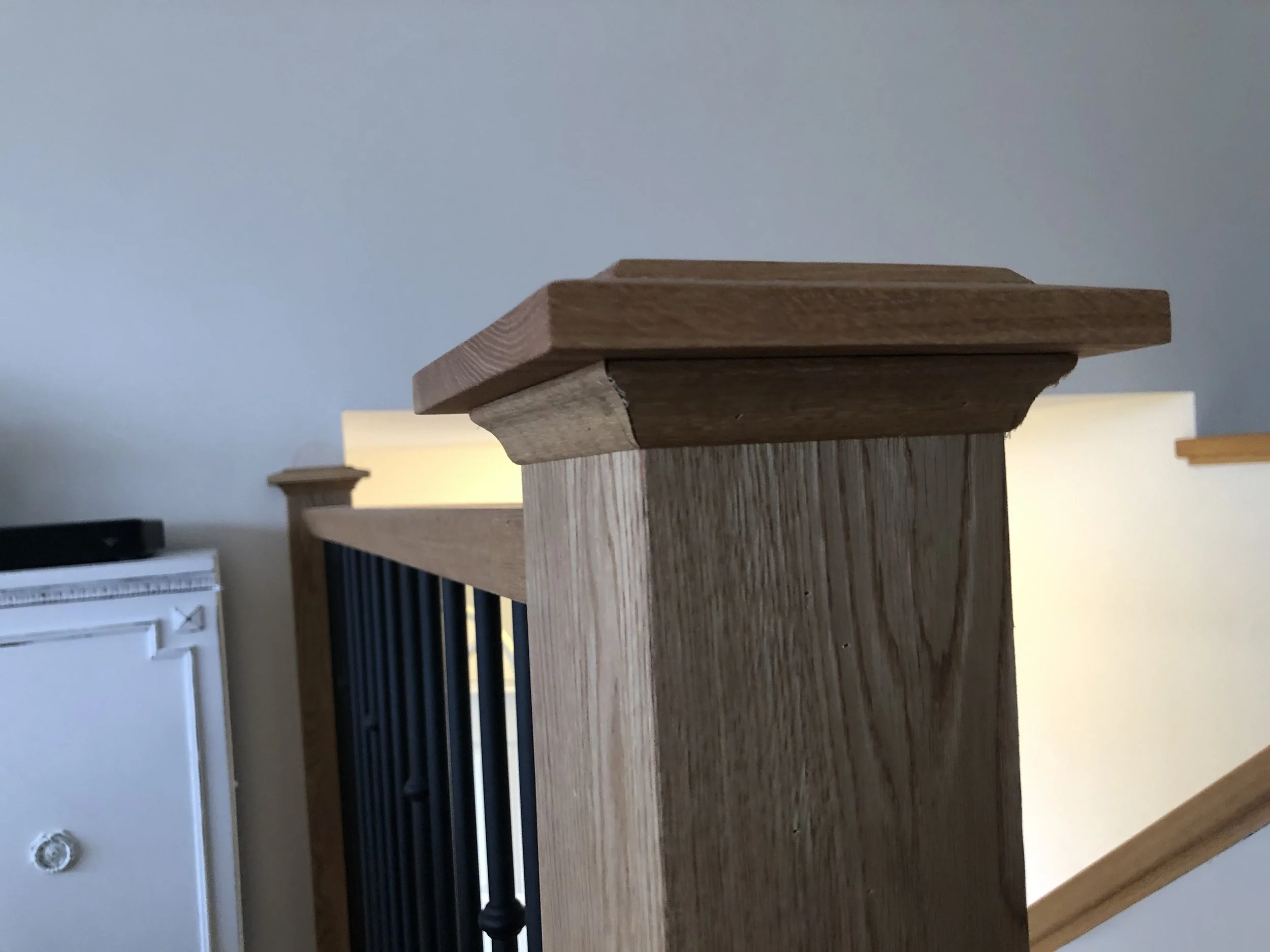 Close-up of a wooden railing or bannister with a decorative molding at the top, set against a painted wall, with some furniture and a window with blinds visible in the background.