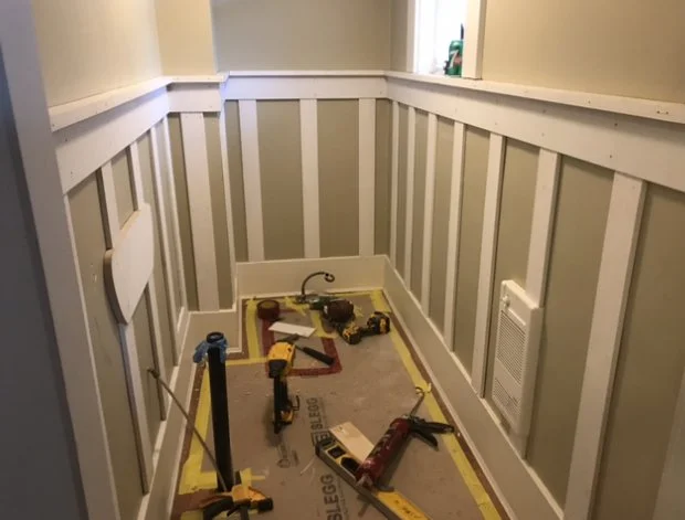 Corner of a room with wainscoting paneling, tools, and supplies on the floor for woodwork or electrical work, including a saw, drill, and measuring tape.