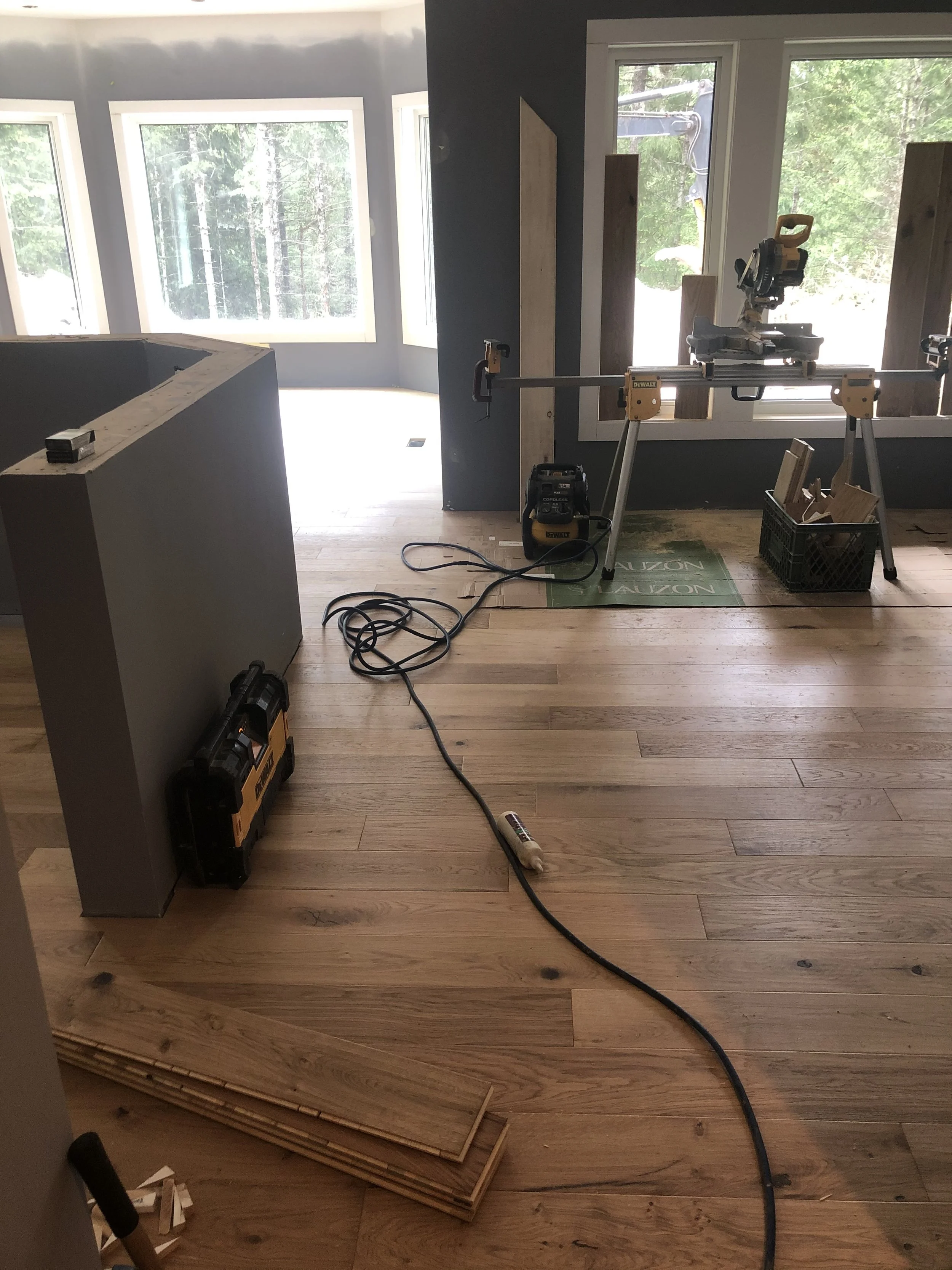 Interior of a room under construction with hardwood flooring, large windows, construction tools, and materials including a miter saw on a stand, wood pieces, a toolbox, and a vacuum cleaner.
