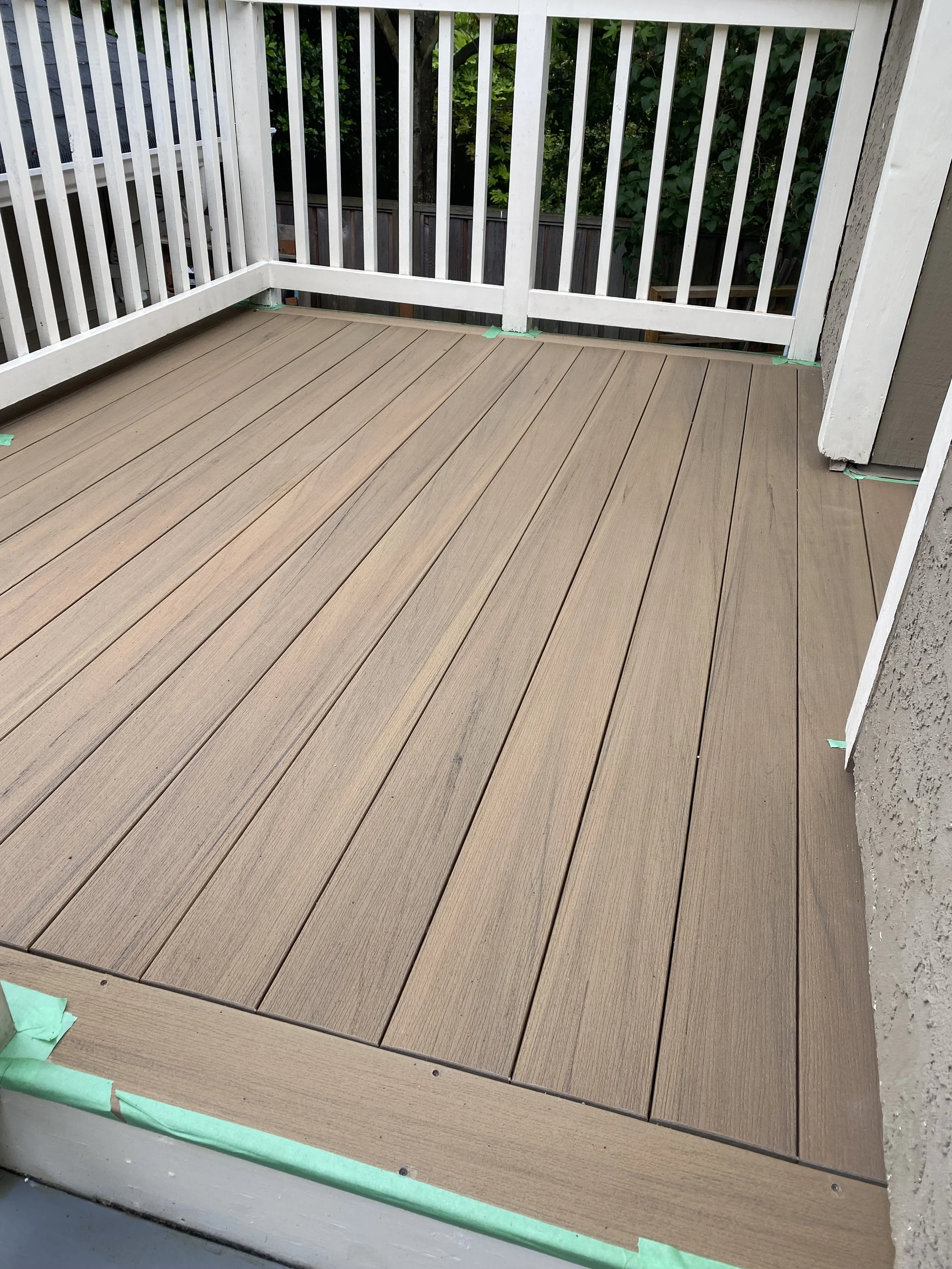 A newly installed wooden deck with light brown planks, partially covered with green painter's tape at the edges, and a white railing surrounding it.