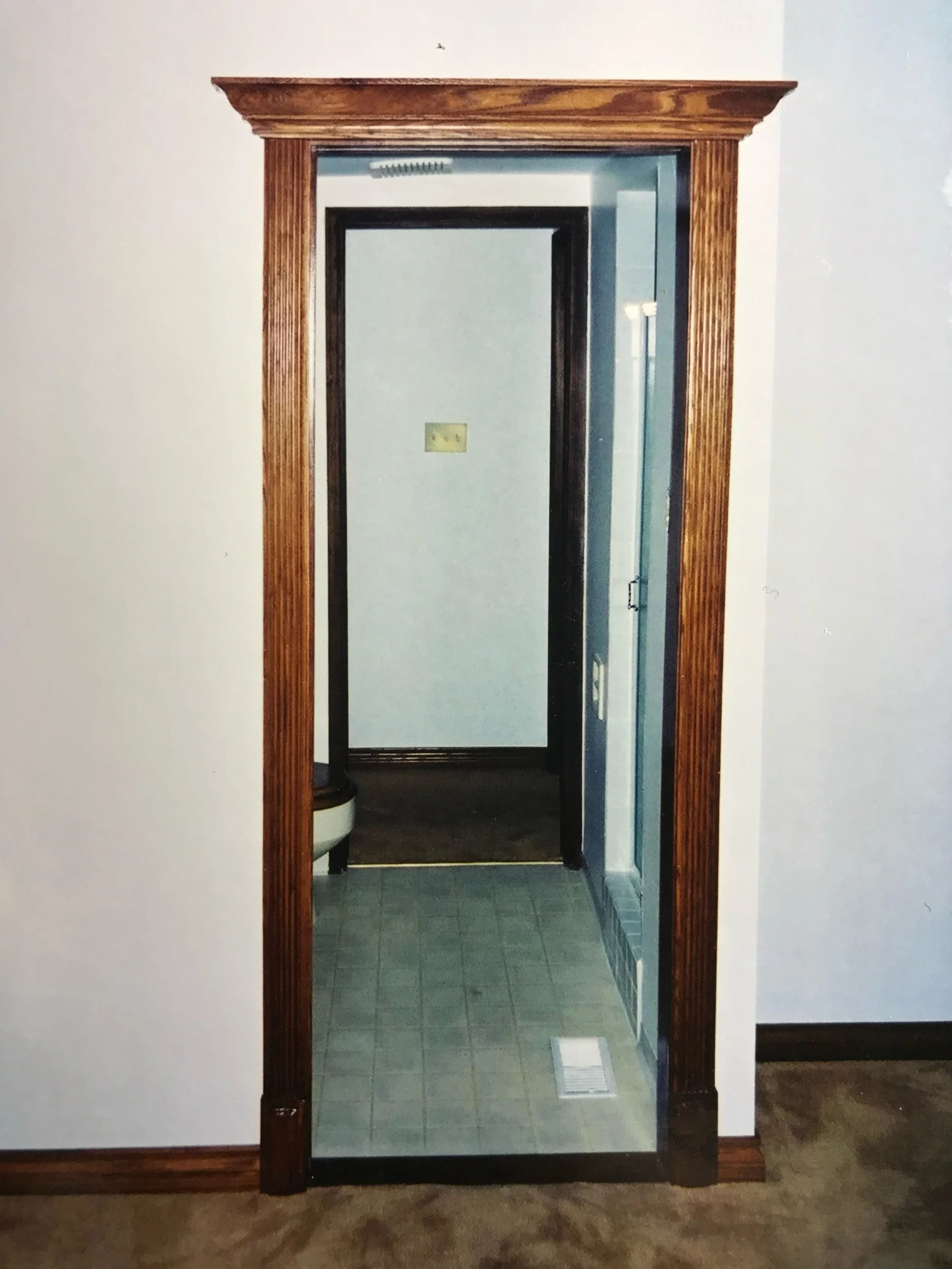 Photo of a doorway with a wooden frame leading into a small room or closet. Inside, there is a mirror on the back wall, a small section of a toilet, and a small window or vent. The floor is tiled with greenish-gray tiles.
