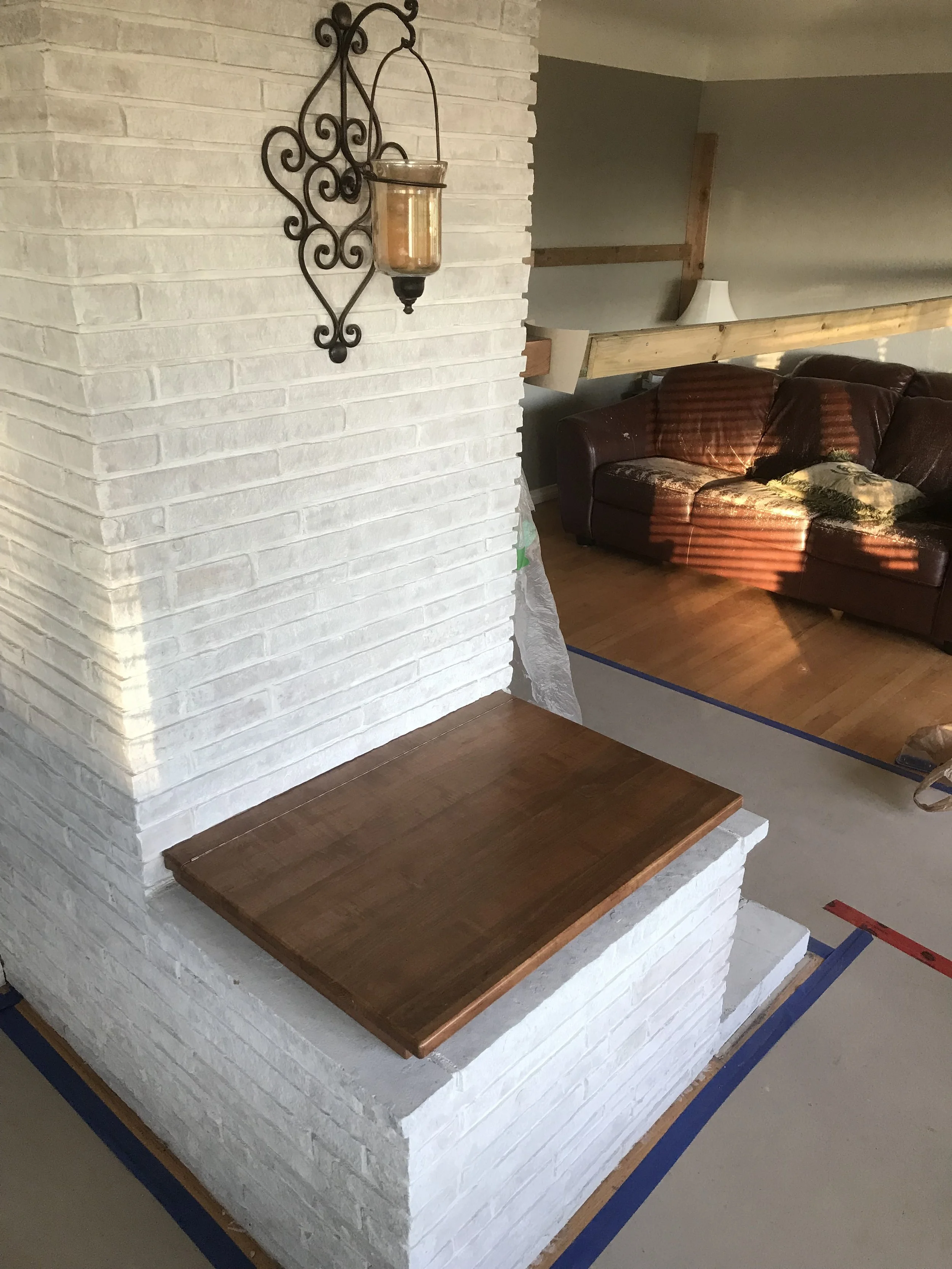 A corner of a living room with a white brick fireplace, a wooden shelf extending from the fireplace, a dark red leather couch with a decorative pillow, a lamp, and a wall with wooden beams and drywall.