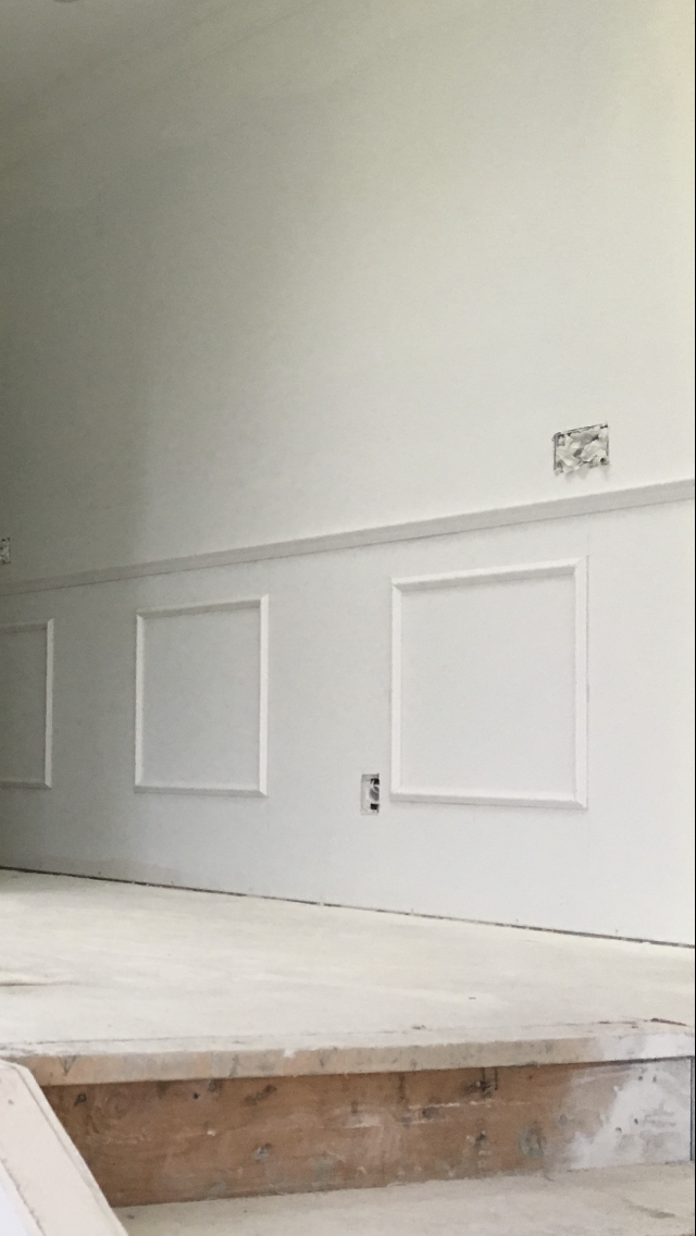 Interior wall under construction with electrical outlets and a cutout for a future fixture, painted in light gray, with wainscoting installed. The floor appears unfinished with exposed wood.