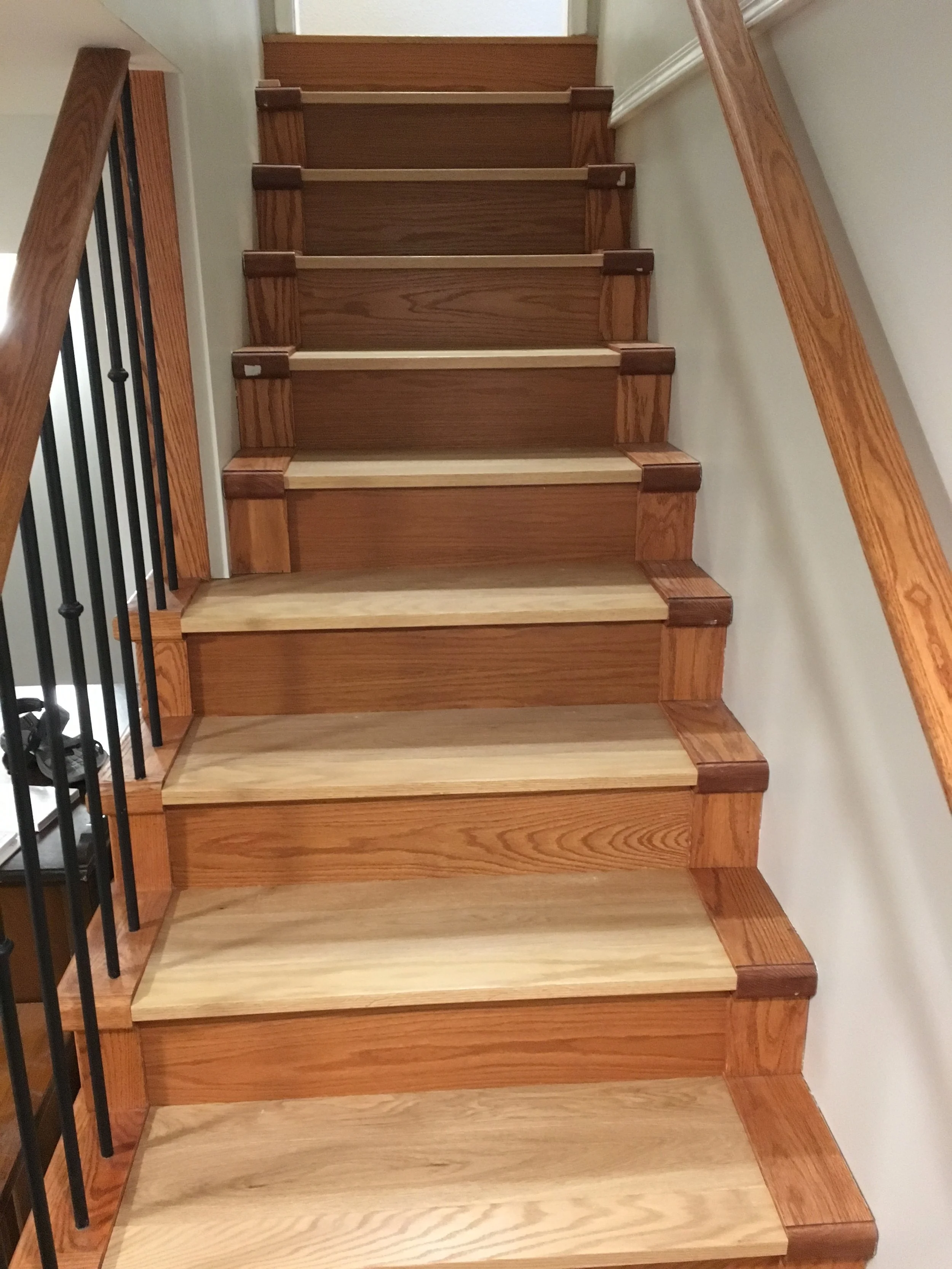 A wooden staircase with blond treads and darker reddish-brown risers, bordered by black metal balusters on the left and a wooden handrail on the right, leading up to a closed white door.