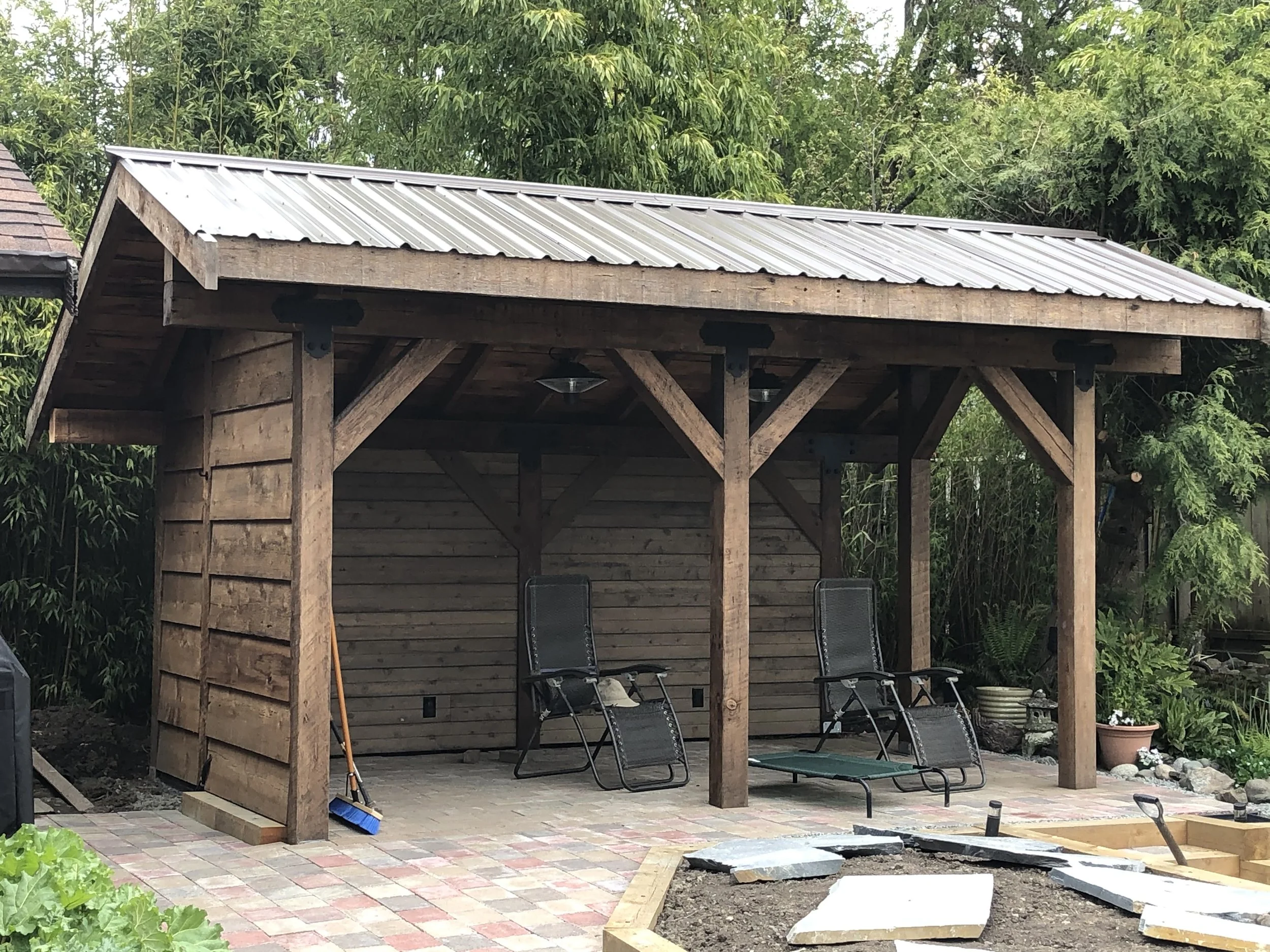 A wooden outdoor shelter with a metal roof, two black lounge chairs underneath, set on a paved patio, surrounded by green trees and plants.