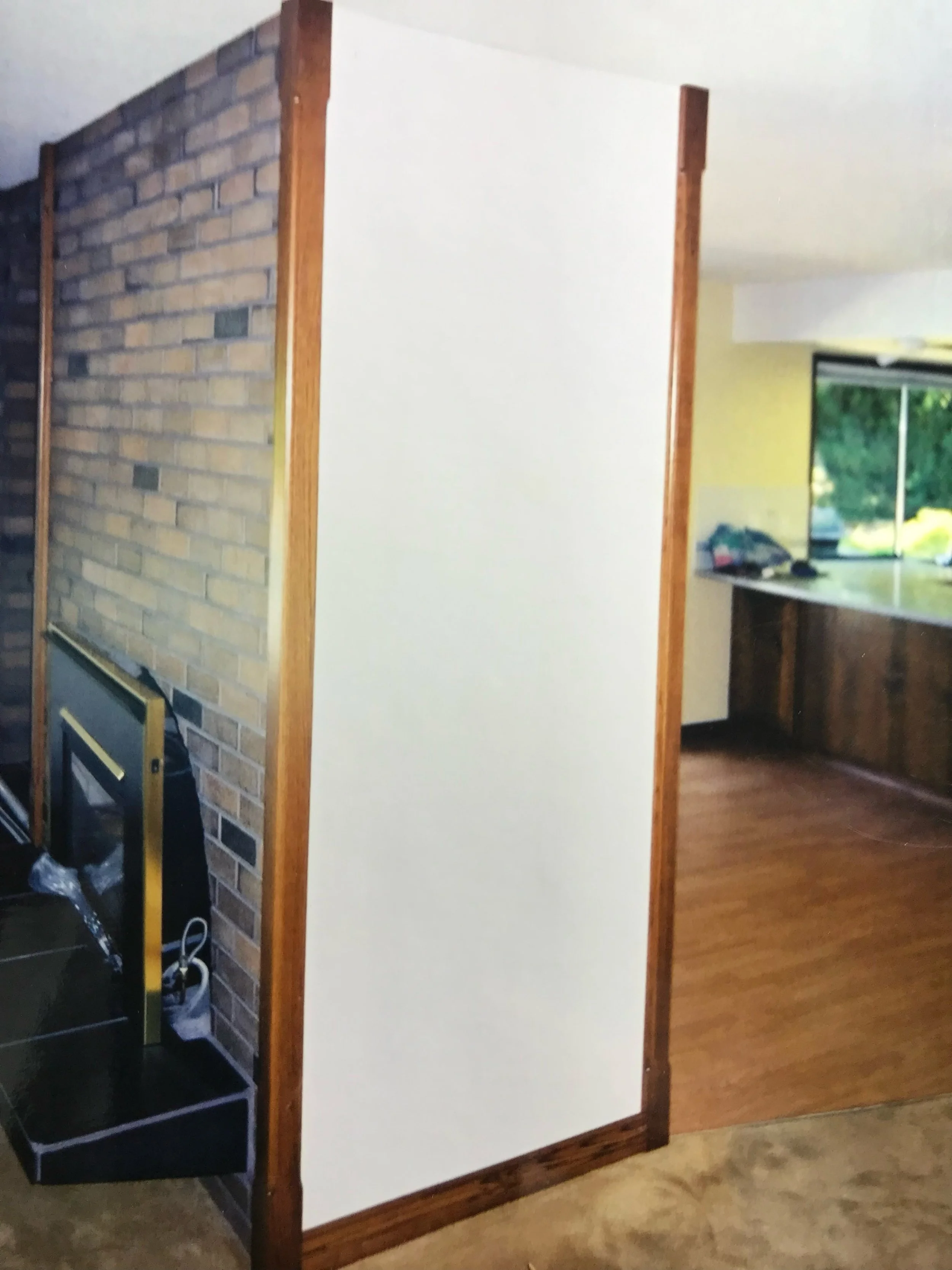 A room with a brick fireplace on the left side, a white or cream-colored wall in the middle, wooden trim around the walls, and a window showing greenery outside in the background.