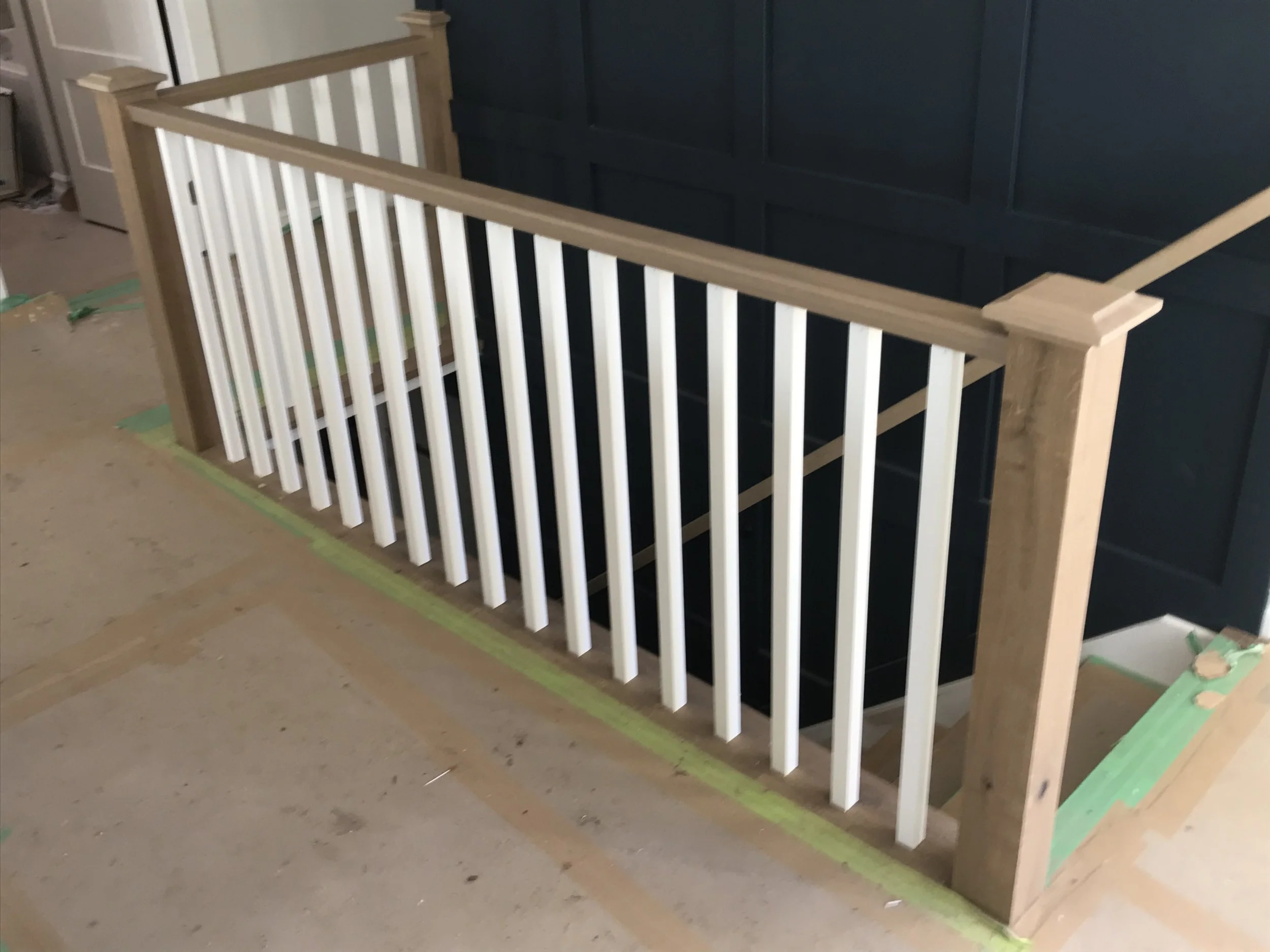 Partial view of a staircase with wooden handrails and white balusters in a construction or renovation setting, with painted and taped floors.
