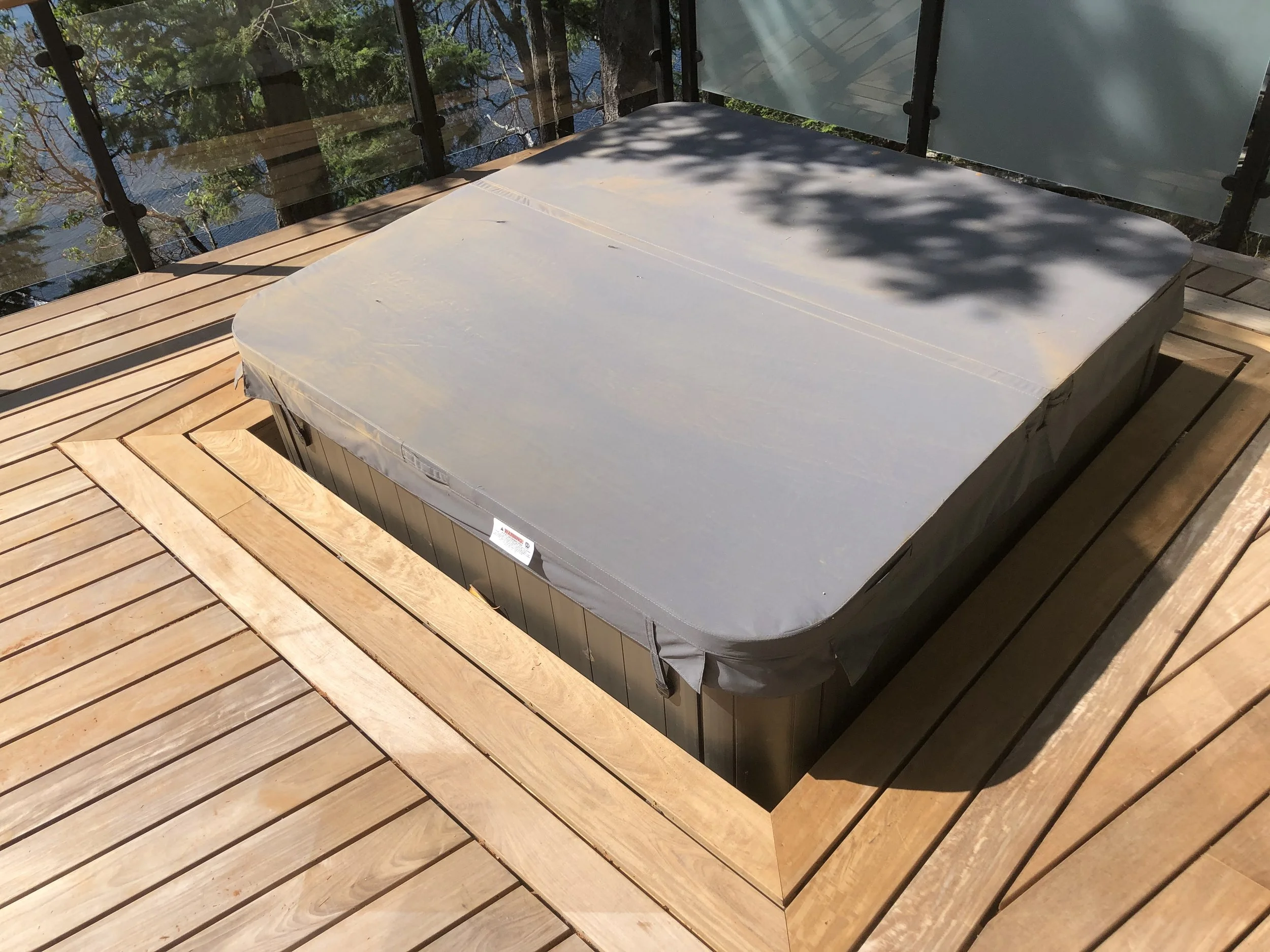 View of a hot tub covered with a protective cover situated on a wooden deck, with a view of trees and water in the background.