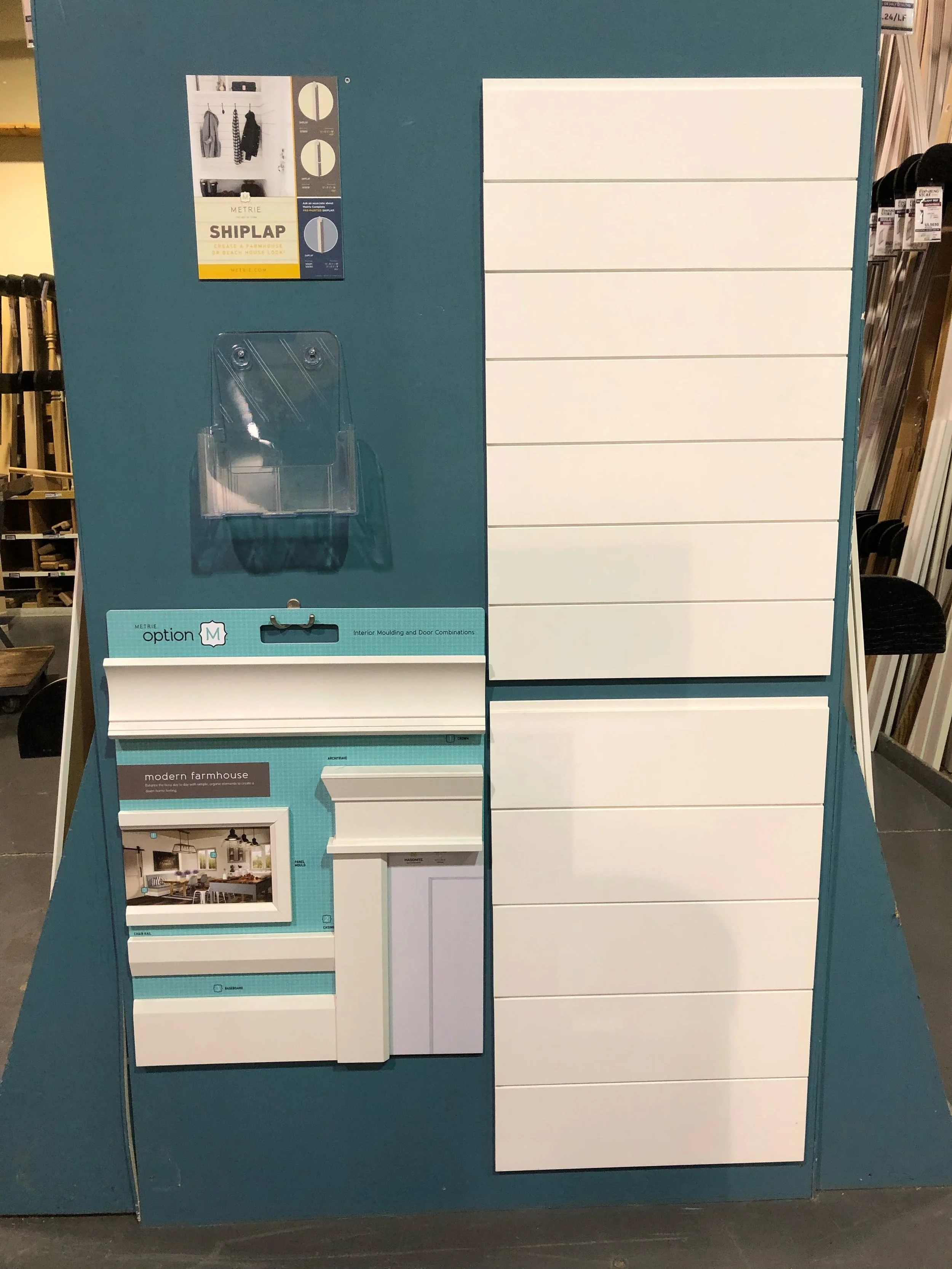 Display board with various interior design samples including white cabinet doors, a sample of a wall-mounted shelf, a magazine or brochure showing a modern farmhouse kitchen, and sample knobs and handles.
