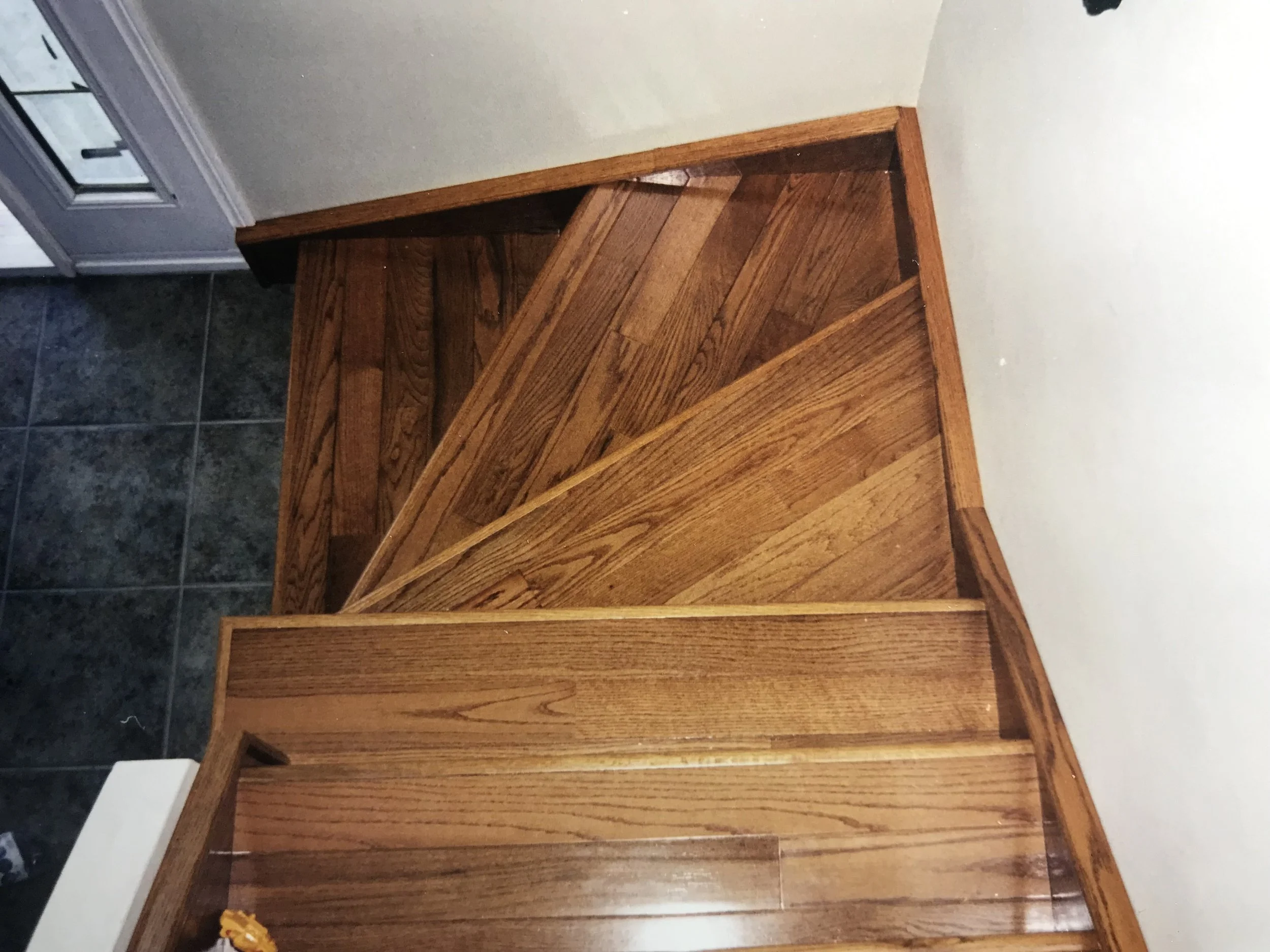 A wooden staircase with triangular panels, finished in a natural wood stain, leading to an upper level near a white wall. A portion of dark tile flooring is visible at the bottom left. There's a window on the left side of the image.