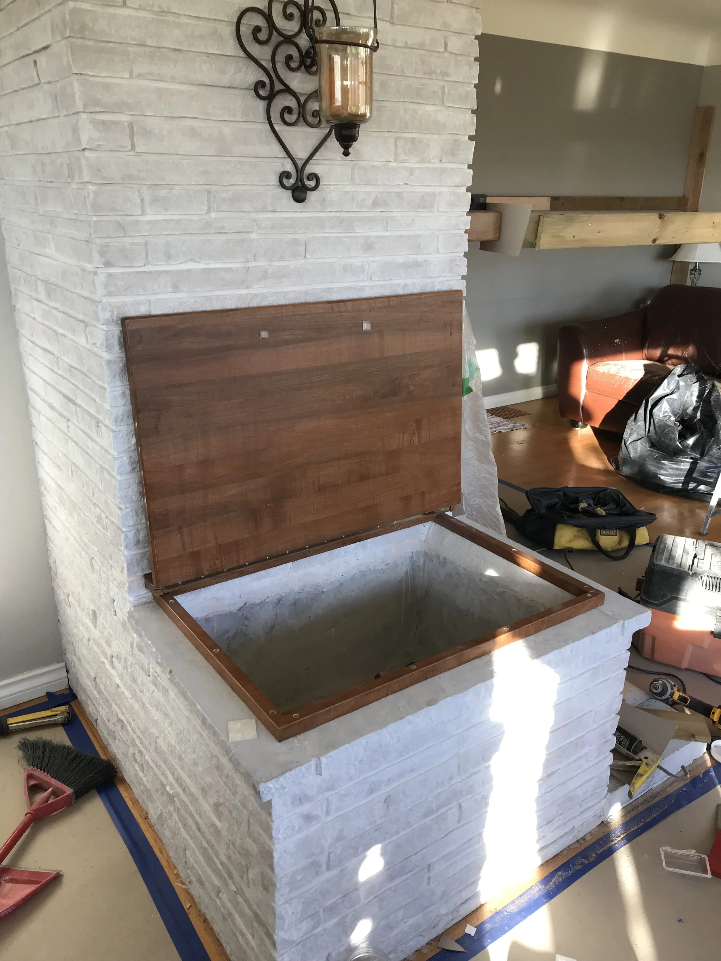 Interior of a house under construction, showing a white brick fireplace with a wooden mantel, and construction tools and materials scattered around.