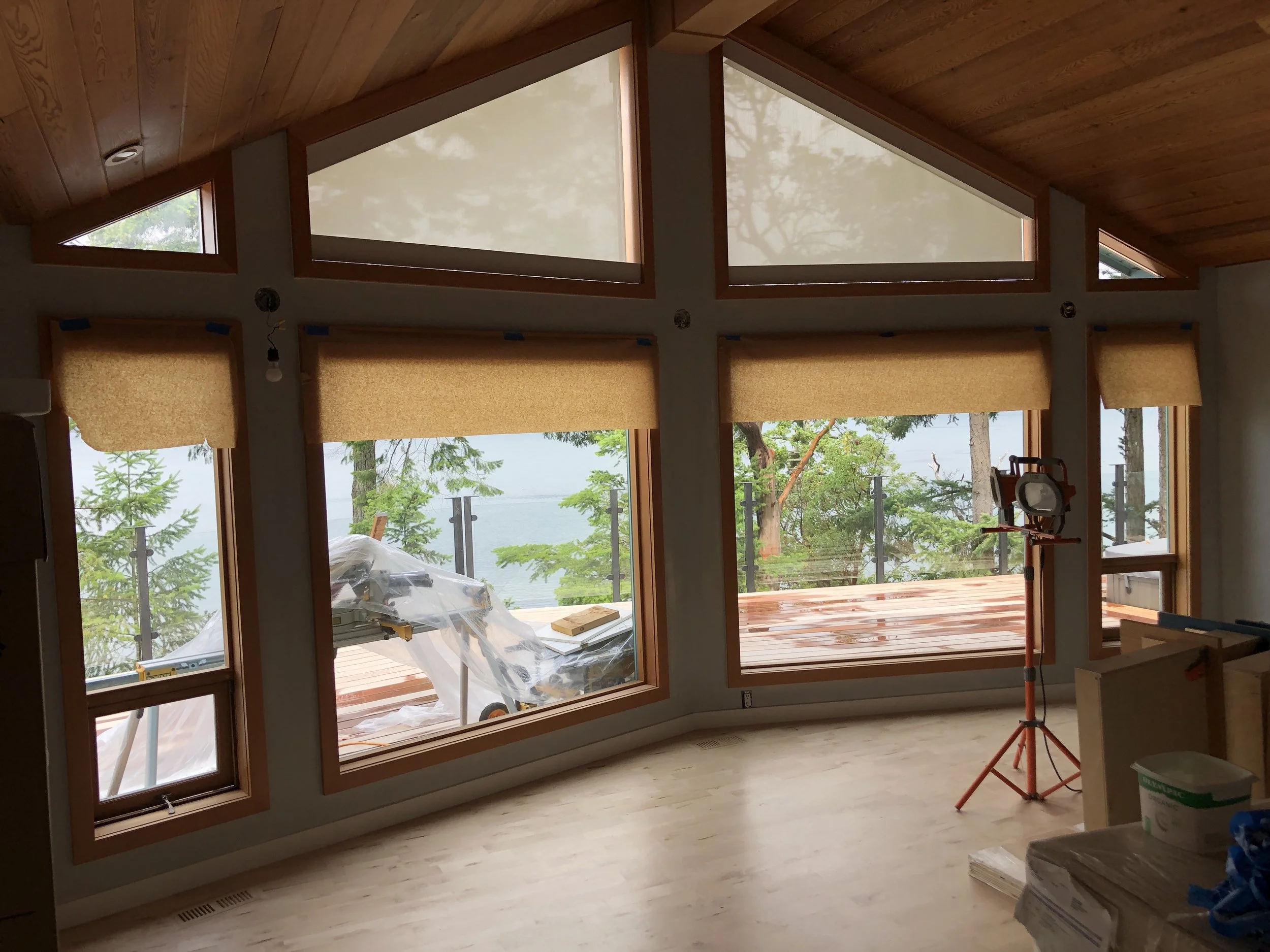 Interior of a house under construction with large windows, wood ceiling, and a view of trees outside.