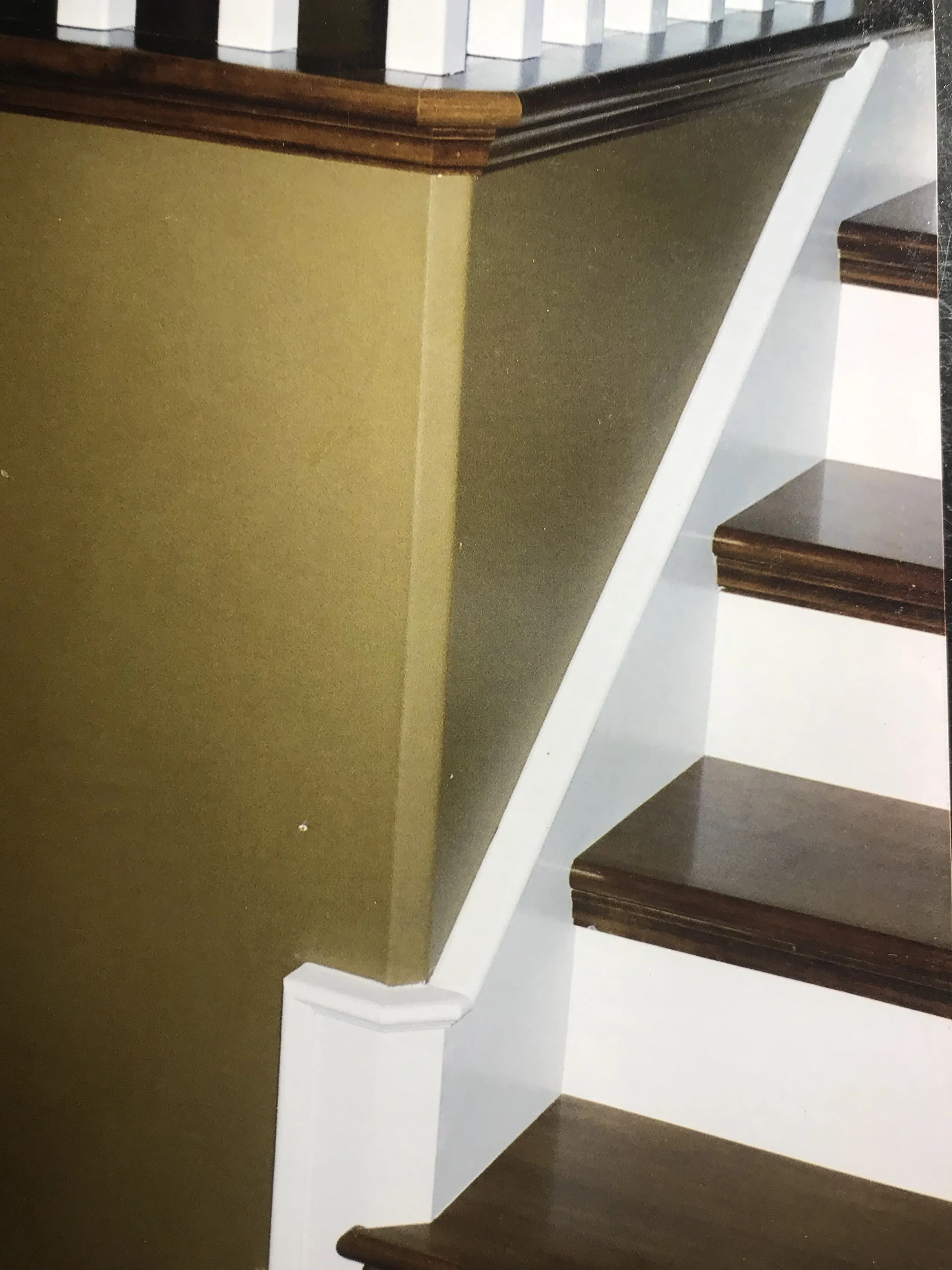 Close-up view of a staircase with dark wooden steps, white risers, and a white trim alongside a partial view of a green wall and a beige wall, with a wooden handrail on top of the green wall.