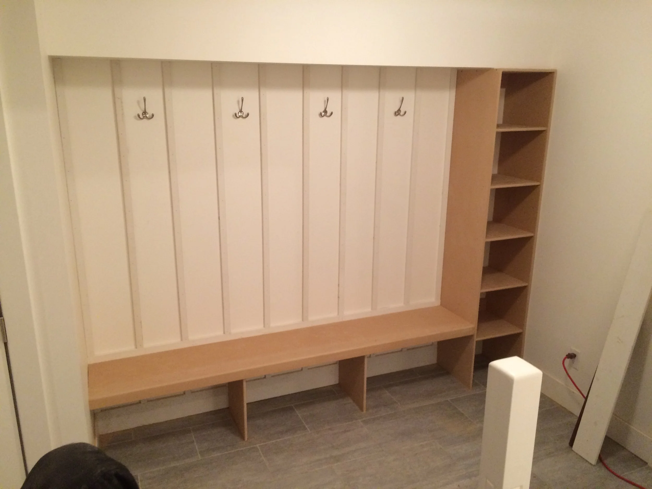 Empty wooden storage unit with hooks on a white wall and open shelves, situated on tiled flooring in a room.
