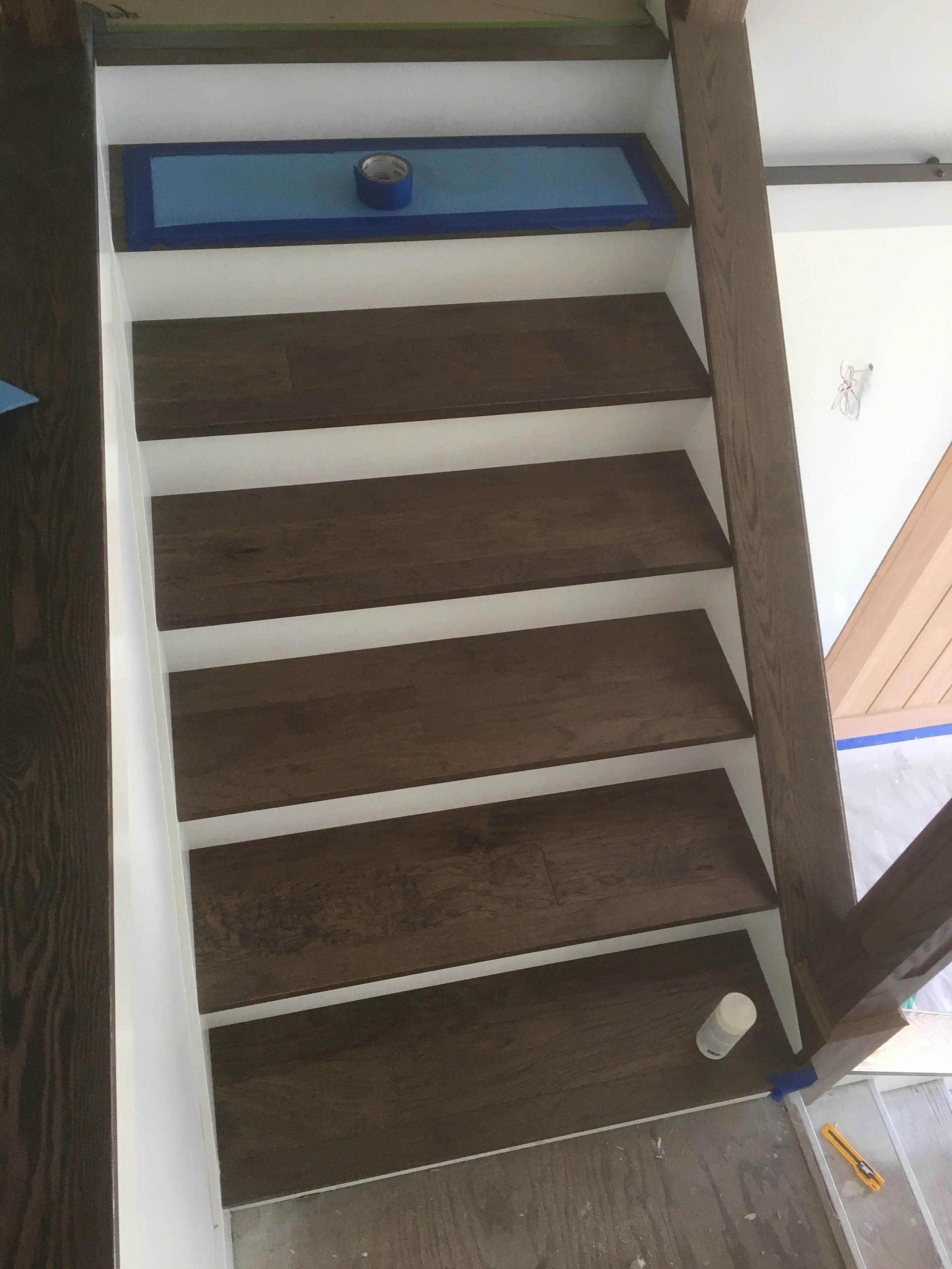 A partially assembled wooden shelving unit with dark brown shelves and white vertical supports, placed on a wooden floor with construction tools nearby, including a cordless drill and a utility knife.