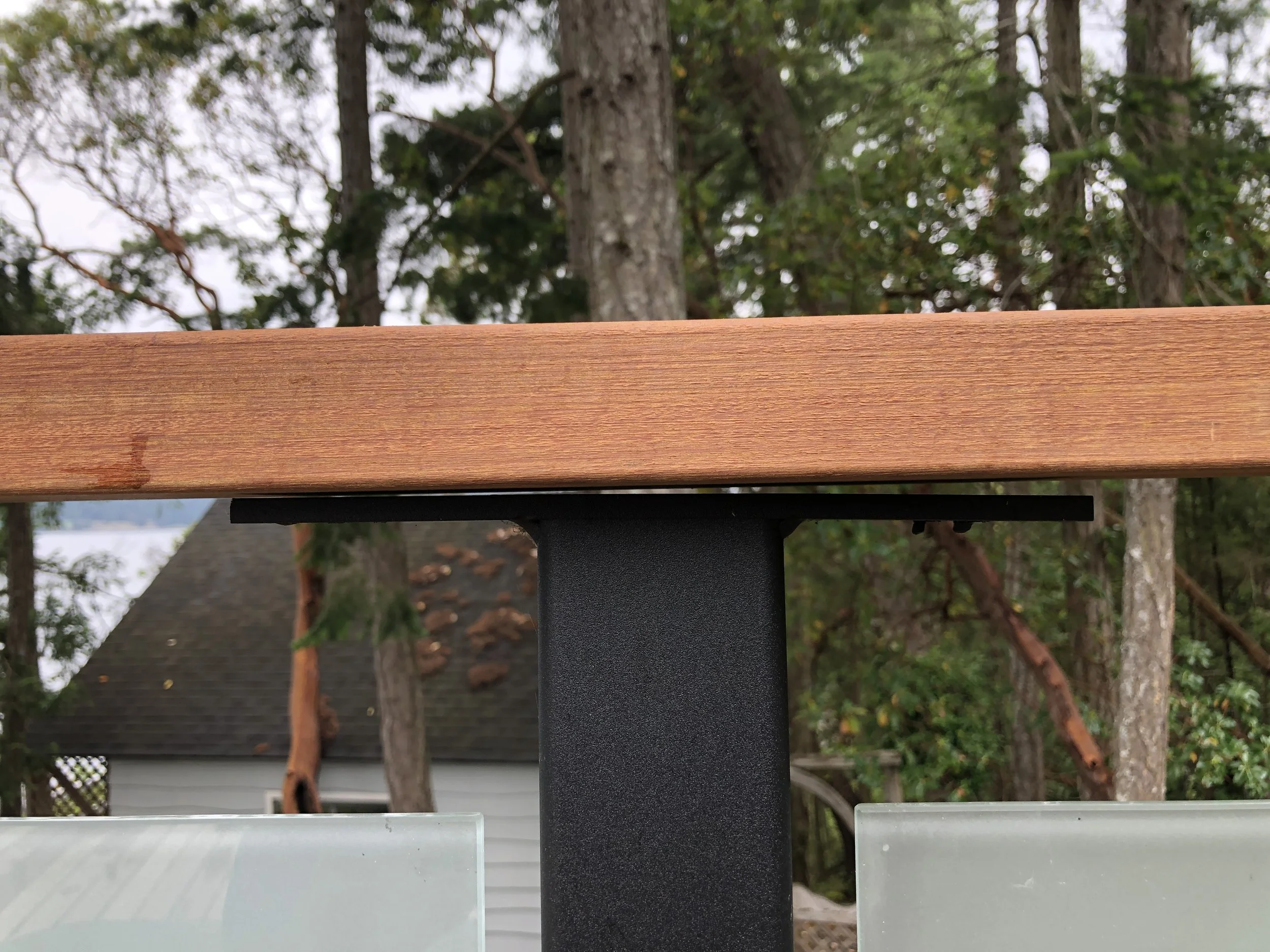 Close-up of a wooden mailbox post with a black metal support, with a backdrop of trees and houses, and frosted glass panels at the bottom.
